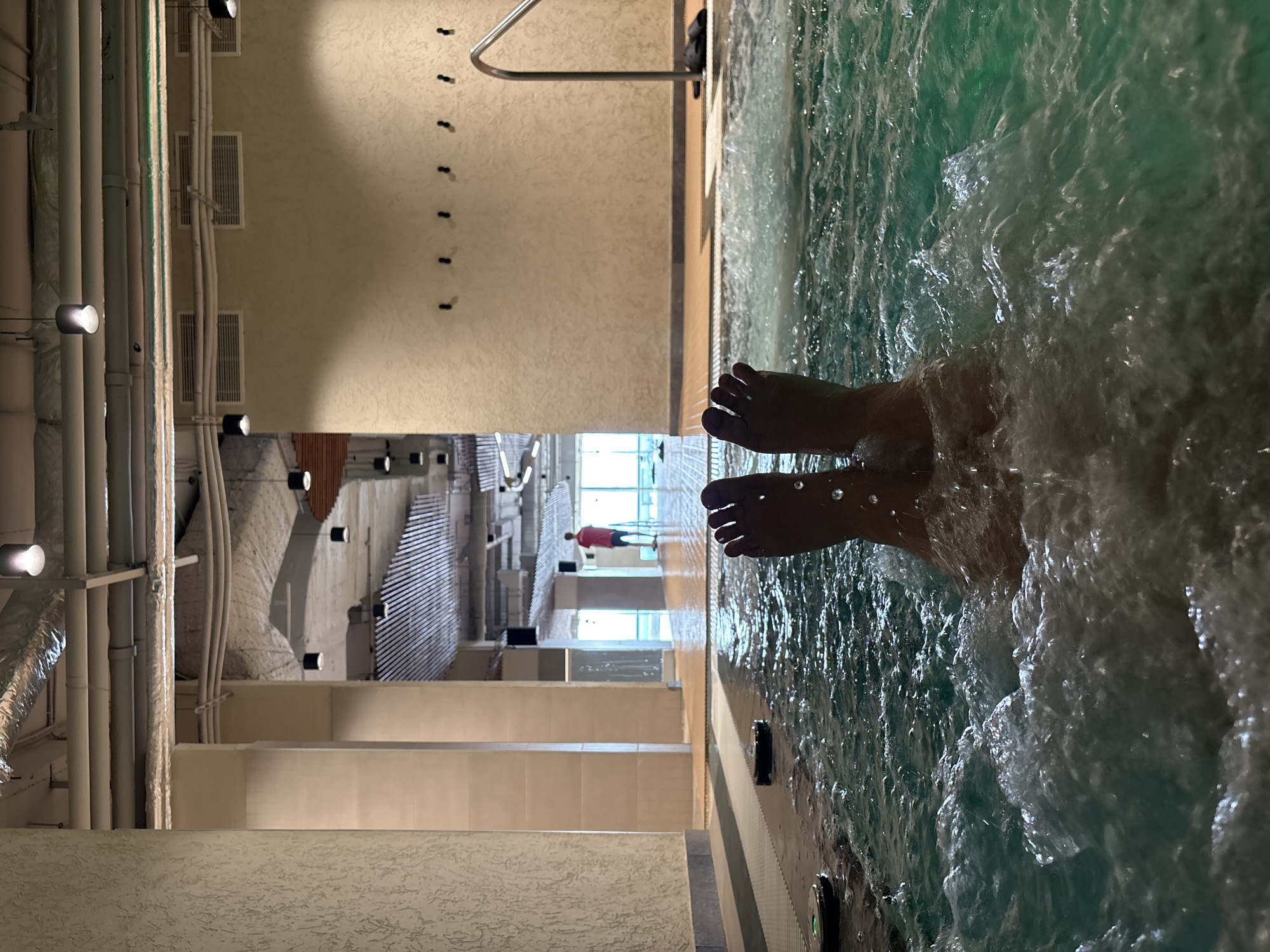 The image shows a person's feet submerged in a hot tub filled with churning turquoise water. The perspective is from within the tub, looking out towards a wider area that appears to be a spa or pool facility. The feet are positioned centrally, with water splashing around them. In the background, a person in a red shirt stands near large windows, seemingly looking out. The environment features industrial-style ceiling elements with exposed pipes and ductwork, along with modern light fixtures. The walls are textured, and there are hooks on one wall. The floor in the background transitions to wooden planks. The location is specified as Khabarovsk, Russia.