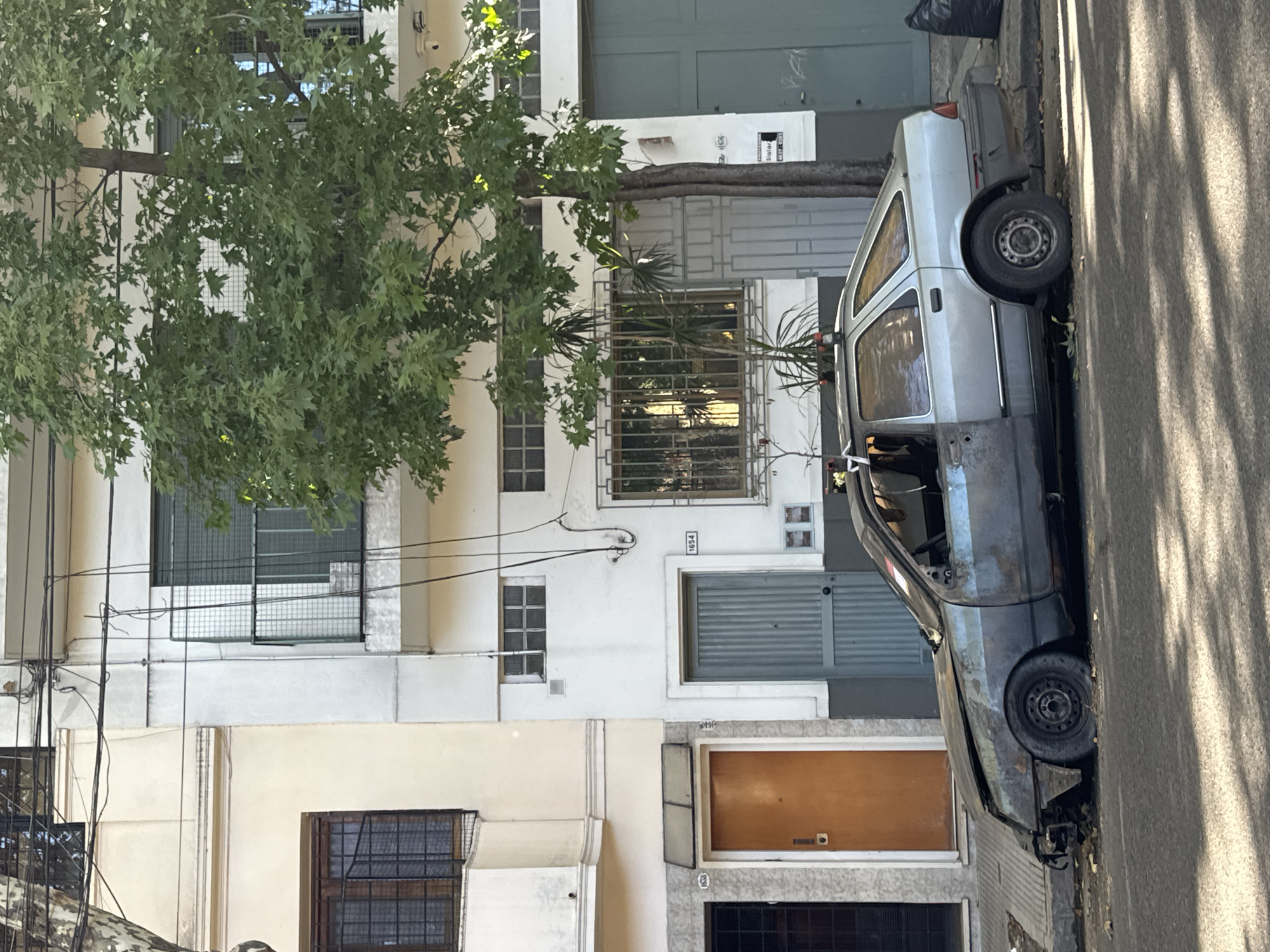 This image captures a residential street scene in Buenos Aires, Argentina, under bright daylight. On the left, a row of multi-story buildings with white and cream-colored facades line the street. Many windows are secured with metal grates, and various doors are visible, including a light blue corrugated garage door, a brown wooden door, and a darker garage door further down. Building numbers "1654" and "1638" are clearly visible on two different structures, with another partial number "16_ _" also present. Numerous black electrical wires are strung along the building exteriors.
A large, leafy green tree, likely a plane tree with broad, lobed leaves, grows in front of the buildings, its branches extending over the street. To the right, an asphalt street runs parallel to the buildings, bordered by a concrete sidewalk. A light-colored (silver-grey) car, appearing to be an older hatchback or small station wagon, is parked along the curb. The car shows signs of significant wear; its front passenger door is missing or heavily damaged, and there is visible dirt or rust on its lower body. Strong, dappled shadows from the tree are cast across the street and the parked car, indicating intense sunshine. A black trash bag rests on the sidewalk behind the car's rear. There are no people visible, and no active events are taking place.