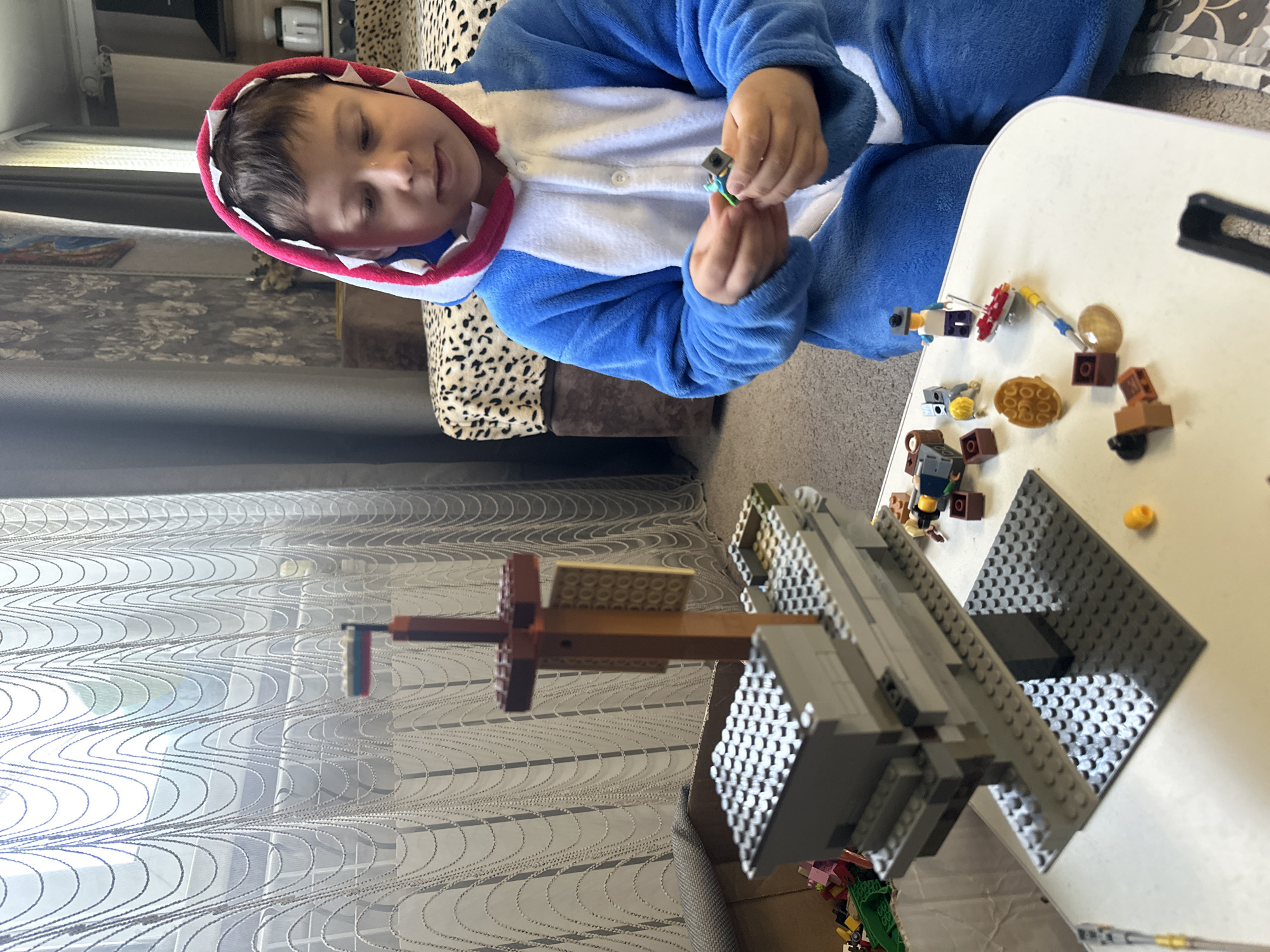 A young child, likely a boy, is depicted sitting indoors, engaged in playing with building blocks. He is wearing a blue and white shark-themed onesie, complete with a hood featuring white teeth-like details around the face. The child is sitting on a sofa upholstered in a distinctive cheetah-print fabric and appears focused on small, colorful building block pieces held in his hands, with a slight smile on his face.
In front of him, on a white table, are numerous other building block pieces of various colors and shapes, including several small figures and accessories. A larger, partially constructed structure made of grey and brown building blocks stands on the table, suggesting an ongoing creation or a completed model. The structure features flat base plates and taller elements, indicating a complex build.
The background reveals a home setting with patterned curtains (light-colored with a wavy design) allowing bright daylight to enter, and a dark grey curtain or blind behind them. A wall with a floral or ornamental wallpaper pattern is also visible. The scene portrays a relaxed, daytime activity, likely in a living room or play area. There is no visible text in the image.
