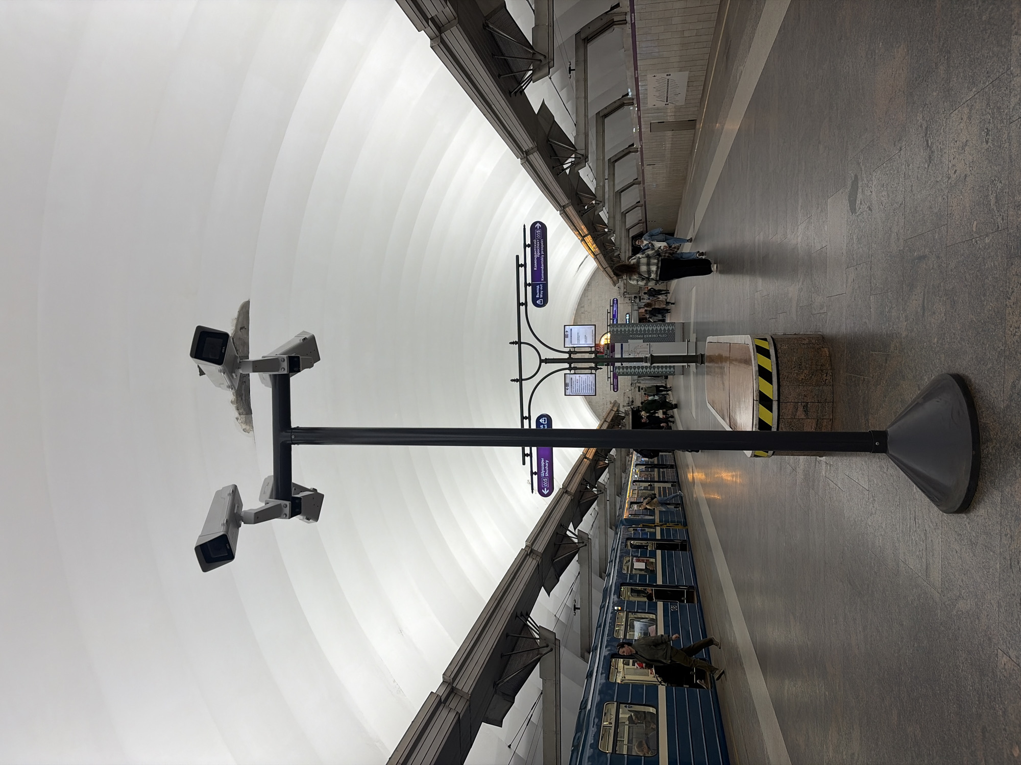 A slightly distorted wide-angle view inside a metro station in Shushary, Russia, featuring a curved white ceiling, a grey granite platform, and a blue and white train. In the center foreground, a black metal pole supports three security cameras facing the platform and a larger base. Several purple signs with white text are mounted on a black frame, positioned behind the pole. People are waiting along the platform and boarding the train. The station is well-lit, reflecting off the ceiling.