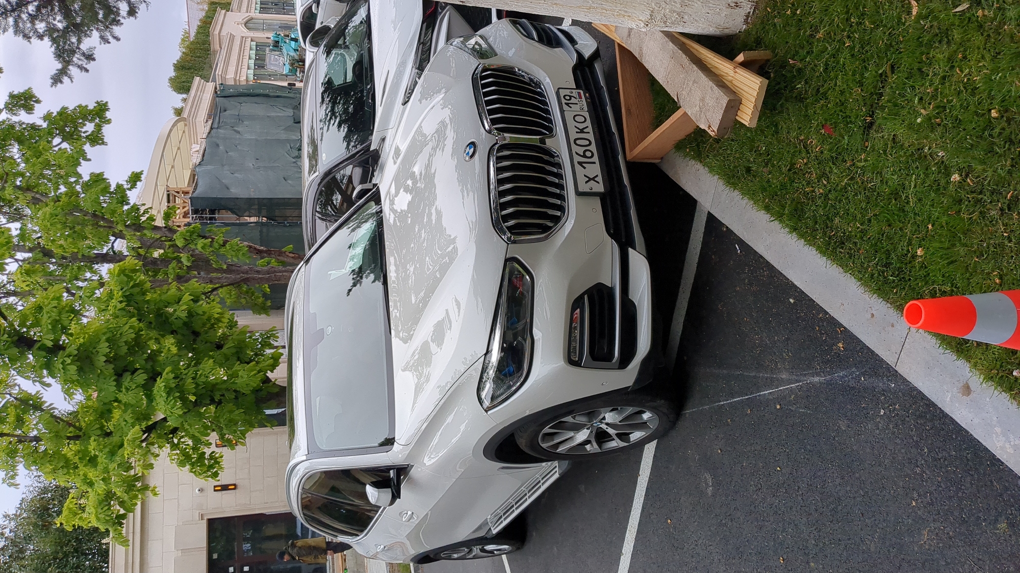 A white BMW X3 is parked on a black asphalt surface in Gelendzhik, Russia, with the license plate X 160 KO 19. The car is next to a curb with green grass and a red and gray traffic cone. Wooden planks lean against the grass near the car. In the background, a building is under construction with scaffolding and green tarps. A green tree is on the left side of the image.