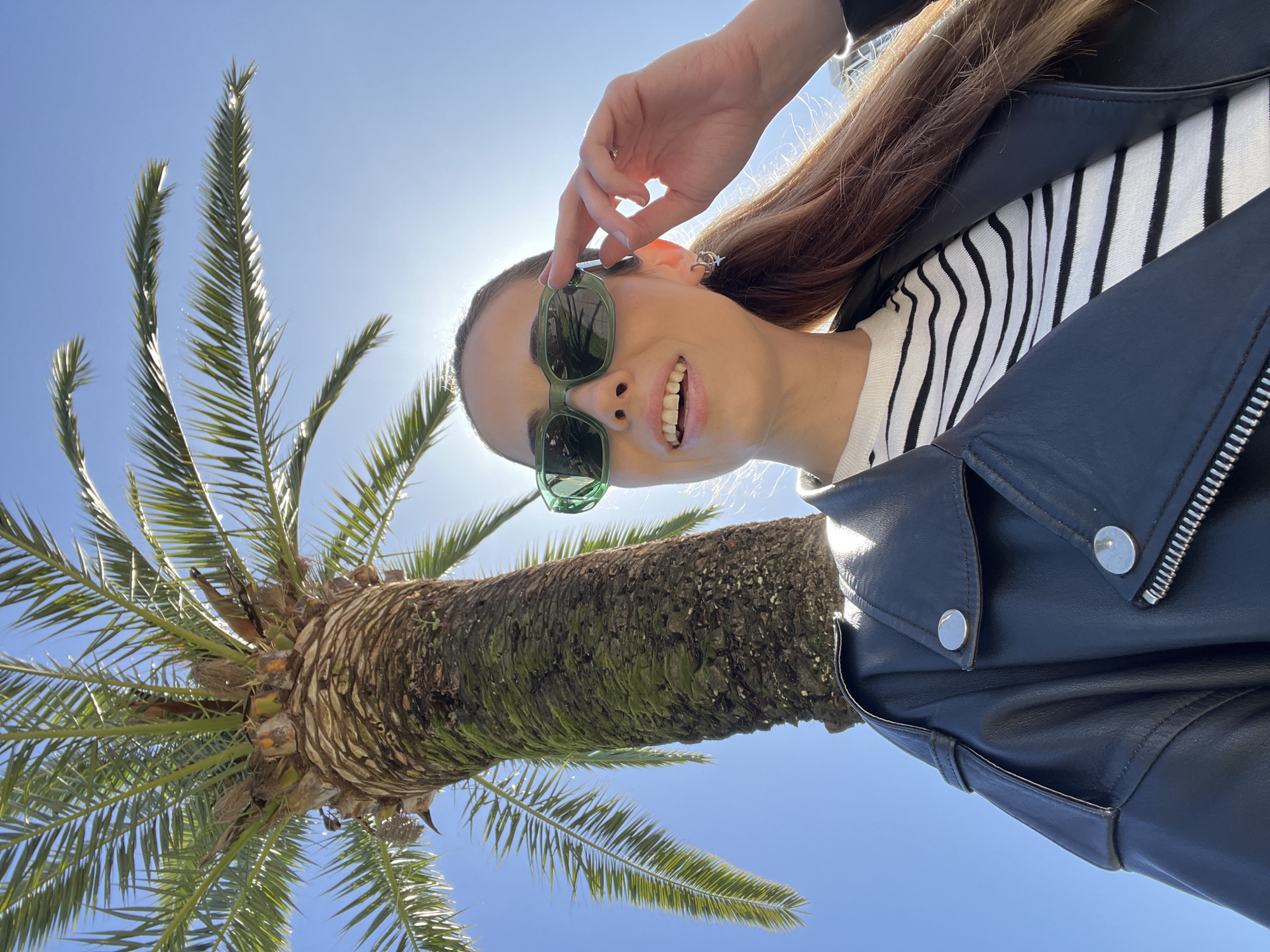 A woman is positioned in the foreground of an image taken from a low angle, with the trunk and fronds of a palm tree prominently featured. The woman is wearing sunglasses and a black leather jacket over a striped top. She has brown hair and is smiling, holding the sunglasses with her left hand. The backdrop is a clear, bright blue sky. The location appears to be in a sunny, outdoor setting, consistent with a coastal city like Bar, Montenegro, where palm trees are common. The tree is in full leaf, and the trunk shows a rough texture. The image captures a moment of relaxation or enjoyment in a warm climate.