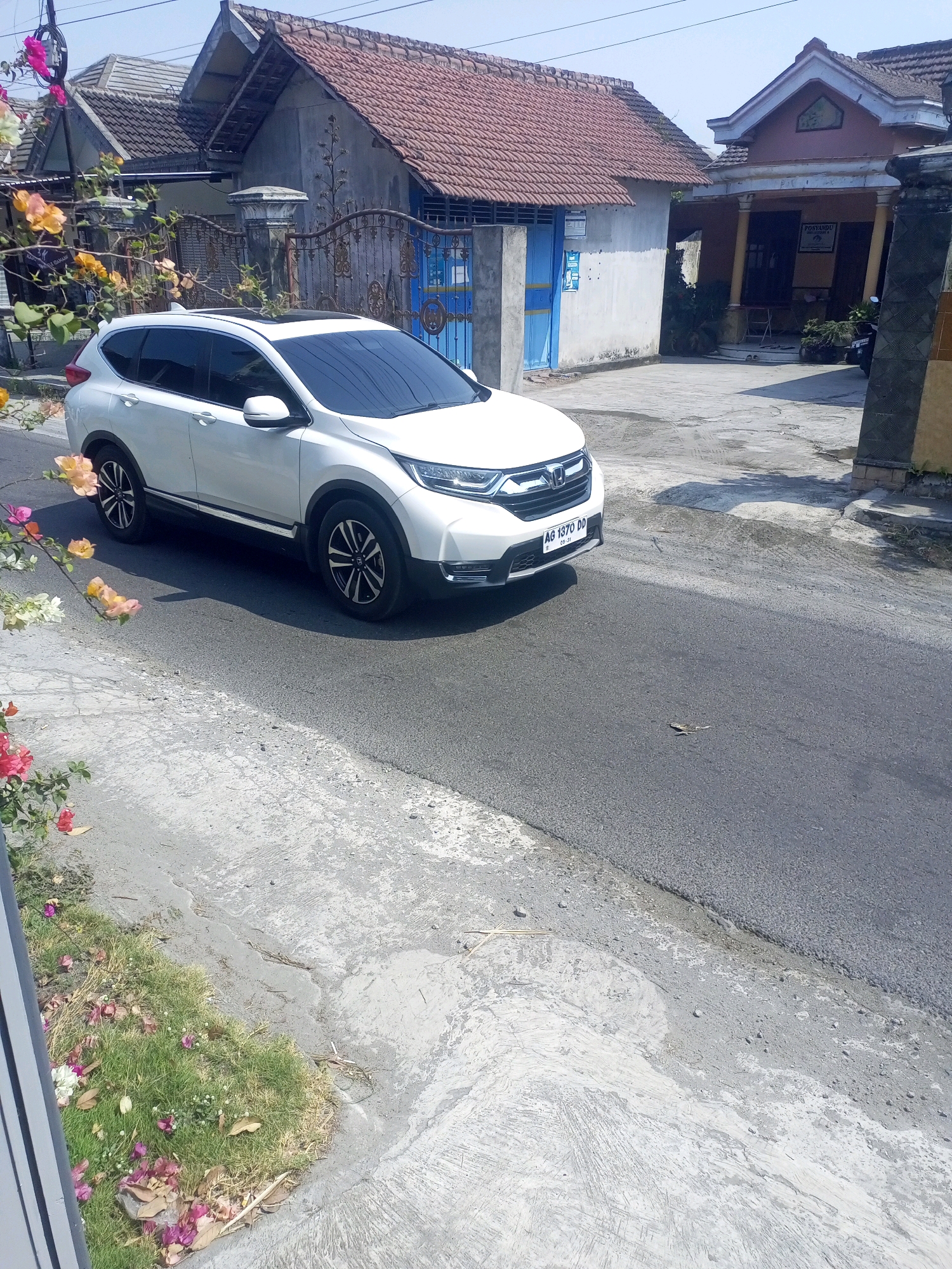 A white Honda CR-V is parked on the side of a street in Ngasem, Indonesia. The car is facing left and is partially obscured by flowering plants. The street is paved and has a concrete gutter. In the background, there are houses with tiled roofs and a blue gate. The sky is clear and blue. The license plate of the car is visible and reads "AG 1370 DD".