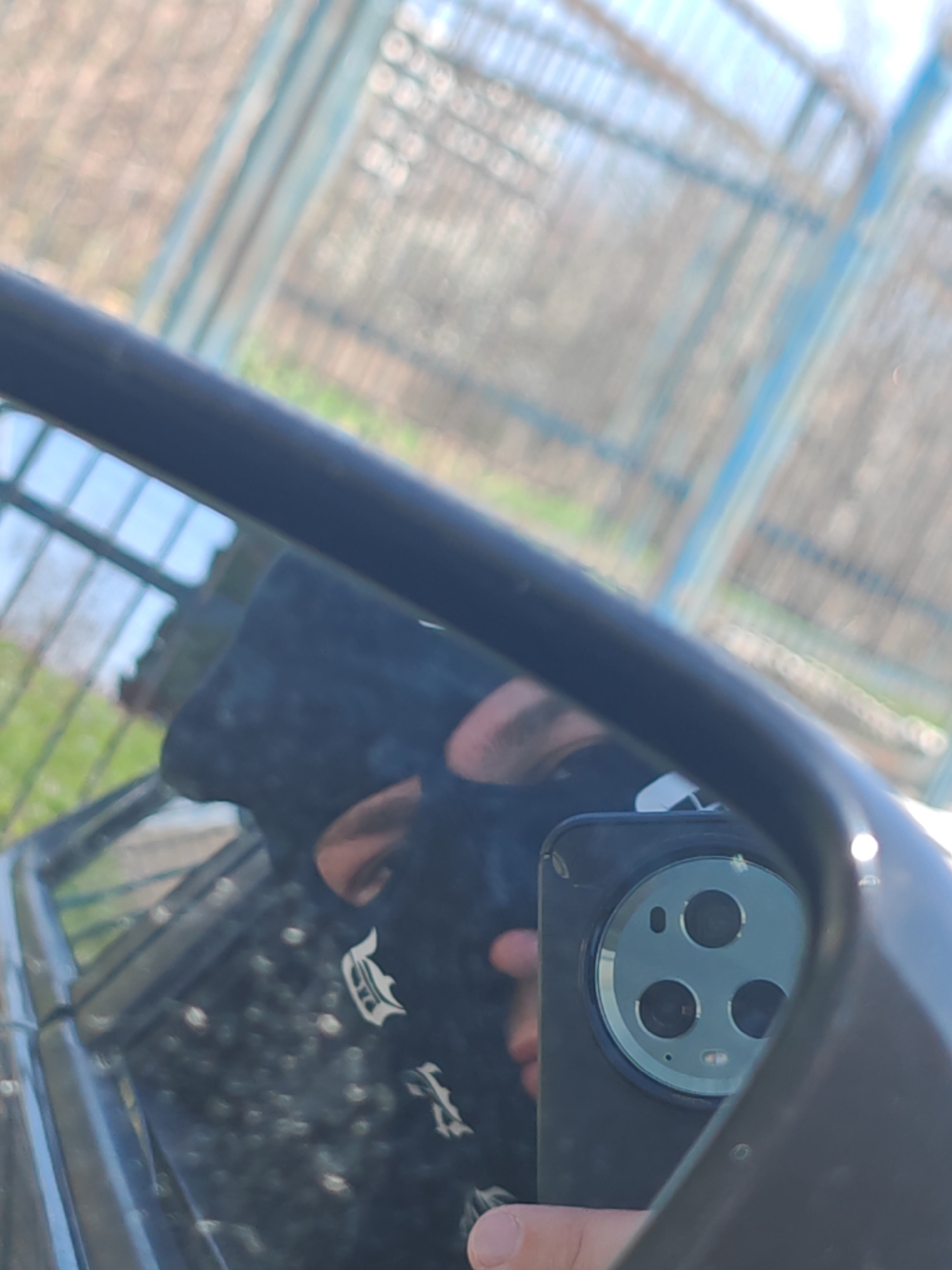 The image displays a reflection within a dark-framed vehicle side mirror during daytime. A light-skinned individual's thumb is visible in the lower-left quadrant, holding a dark-cased smartphone. The reflected smartphone's rear panel features a prominent circular camera module containing three lenses, a flash, and an auxiliary sensor. Portions of the individual's face, specifically an ear and the side of the head, along with a shoulder wearing a dark garment with a white graphic, are also visible in the reflection. The out-of-focus reflected background consists of a light-colored, possibly chain-link, fence with vertical blue supports, green vegetation, and a clear blue sky. No specific location within Shumen, Bulgaria, is identifiable from the provided visual information.