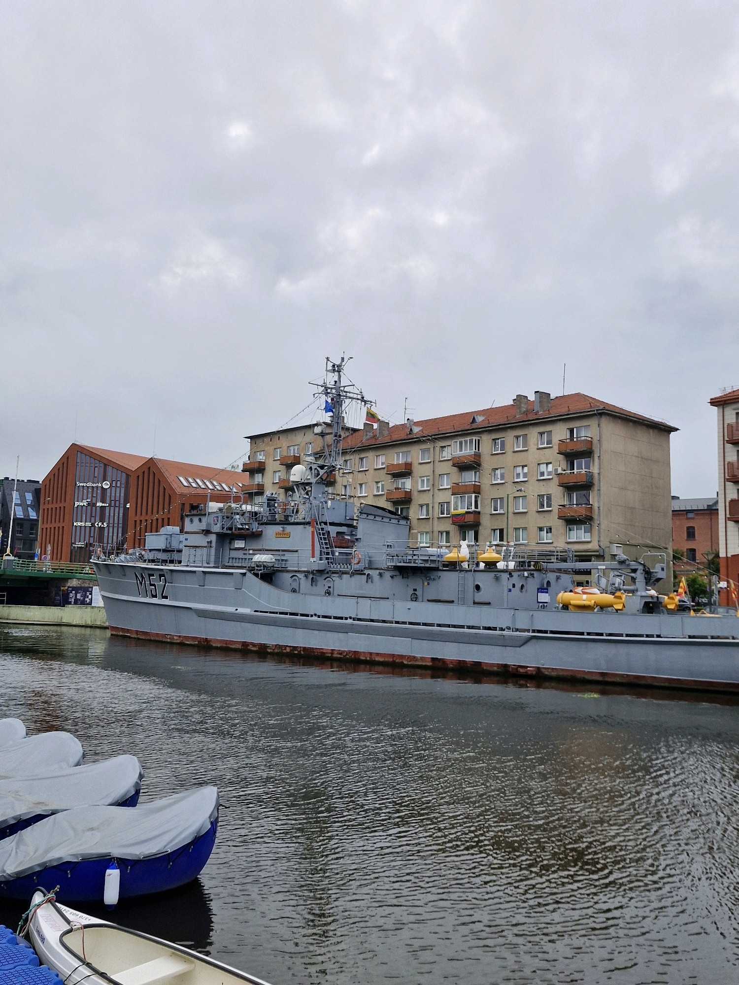 The image depicts a grey naval vessel, marked "M52", docked in a canal or harbor in Klaipėda, Lithuania. The vessel is positioned parallel to the shore, with buildings lining the opposite bank. The architecture of the buildings varies; one is a modern structure with a red, angular roof and glass facade, bearing signage for "Swedbank", "pwc", and "KBS CF.S". Adjacent to this is a multi-story residential building with a light brown facade and numerous balconies. The sky is overcast, contributing to a muted color palette. In the foreground, several small boats, some covered with grey tarps, are moored in the water. The water itself shows ripples, indicating a slight movement. The overall scene suggests a calm, urban waterfront environment.