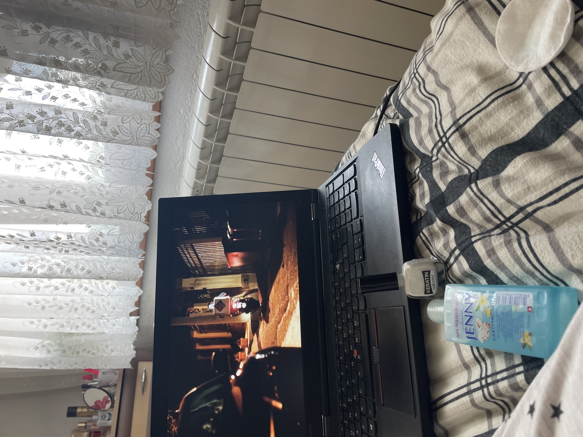 The image depicts an indoor scene, likely a bedroom, with a laptop open on a person's lap, covered by a plaid blanket. The laptop screen displays a nighttime scene featuring a car on the left and a person on a motorcycle with a delivery box on the back to the right. In the foreground, on the blanket, are two bottles of nail polish related products: one is a clear blue bottle labeled "JENNY NAIL POLISH REMOVER" and the other is a small, clear bottle with a white cap labeled "KERATIN nail color." A white cotton pad is visible to the right of the bottles. Behind the laptop, a radiator runs along a textured white wall. Above the radiator, sheer white curtains with a floral pattern are visible, suggesting a window is present. To the far left, on a shelf, are several cosmetic bottles and a small round mirror with a pink floral design, hinting at a vanity or dresser. The overall impression is a relaxed, indoor setting, possibly in Zlatograd, Bulgaria, given the context provided.