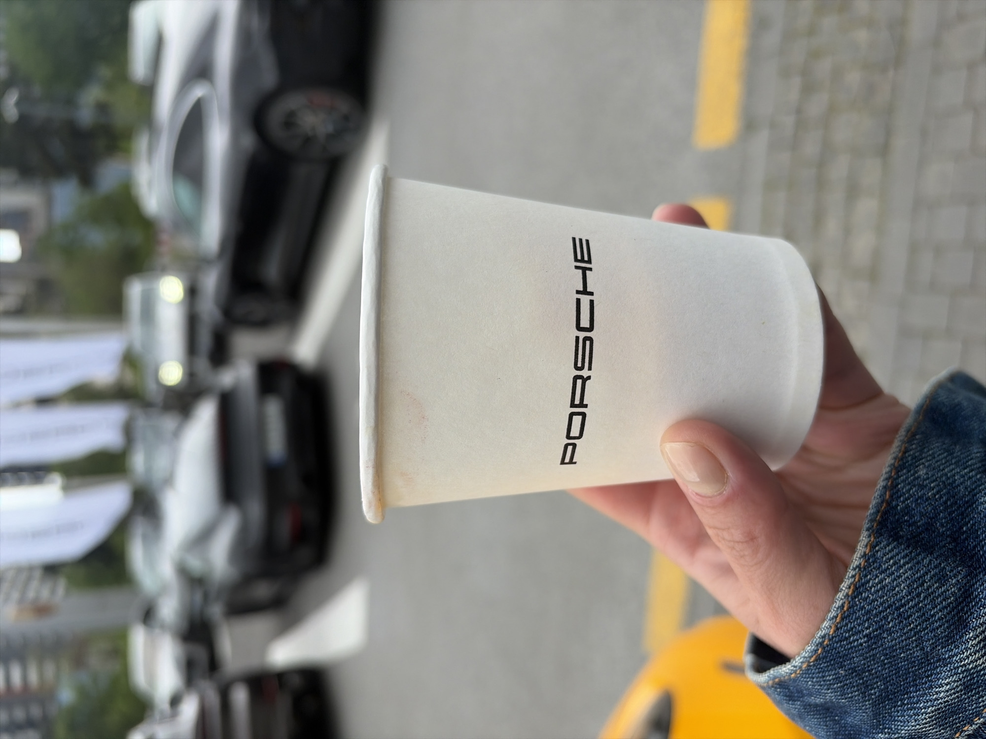 A hand wearing a denim jacket holds a white paper coffee cup with "PORSCHE" printed vertically in black capital letters. In the blurred background, several cars are parked along a street. A yellow curb marking is visible on the right side of the street. The image was likely taken in the city of Tbilisi, Georgia.