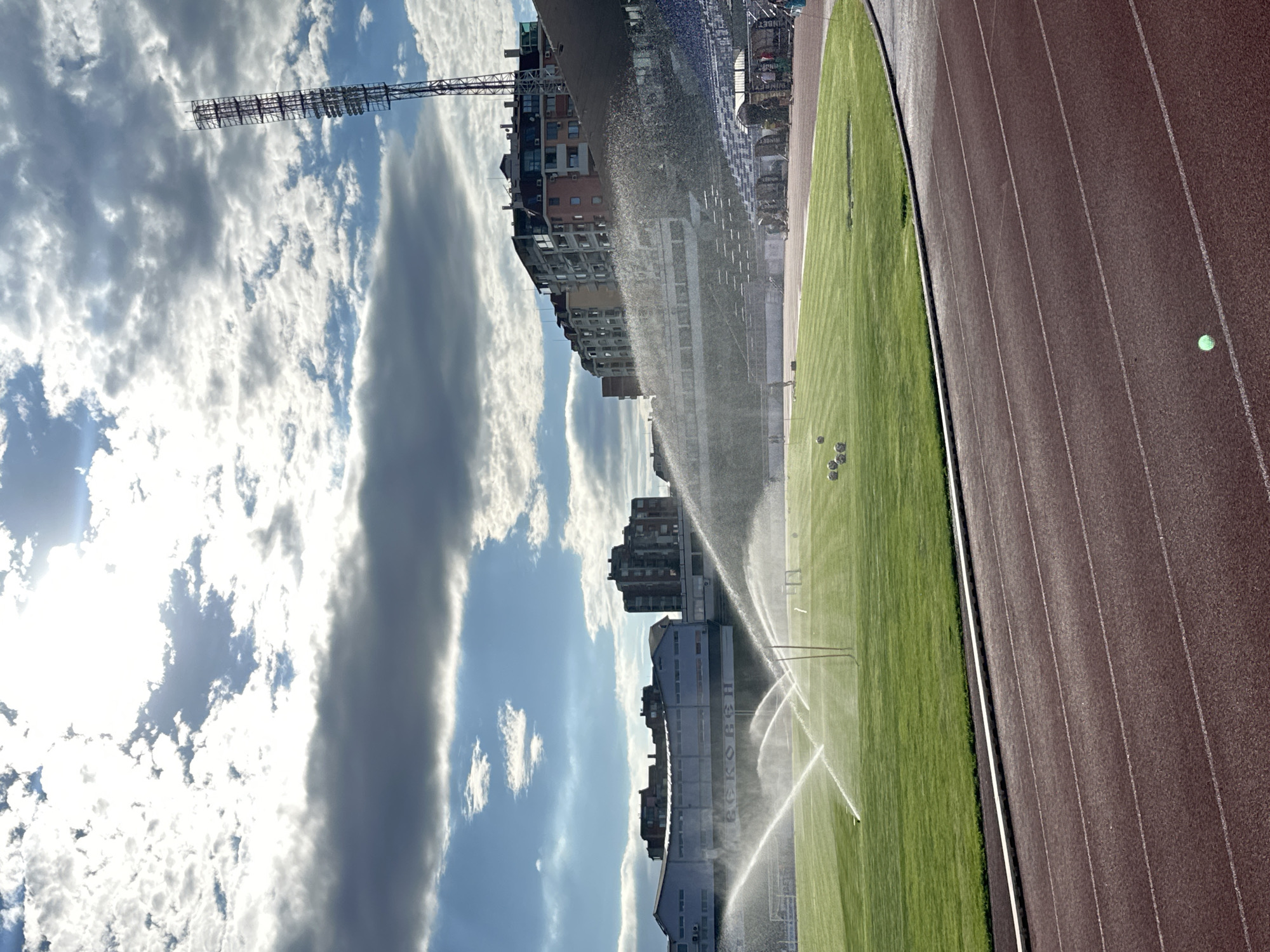 The image shows a stadium in Veliko Tarnovo, Bulgaria, under a cloudy sky. The stadium features a bright green football field with several sprinklers actively watering the grass, creating a misty effect. In the background, multi-story buildings and stadium seating are visible. A tall light tower stands prominently on the right side of the frame. The foreground consists of a red athletic track. Several soccer balls are scattered on the field. A few people can be seen in the distance on the field and in the stands. The sun is partially obscured by clouds, casting rays of light.