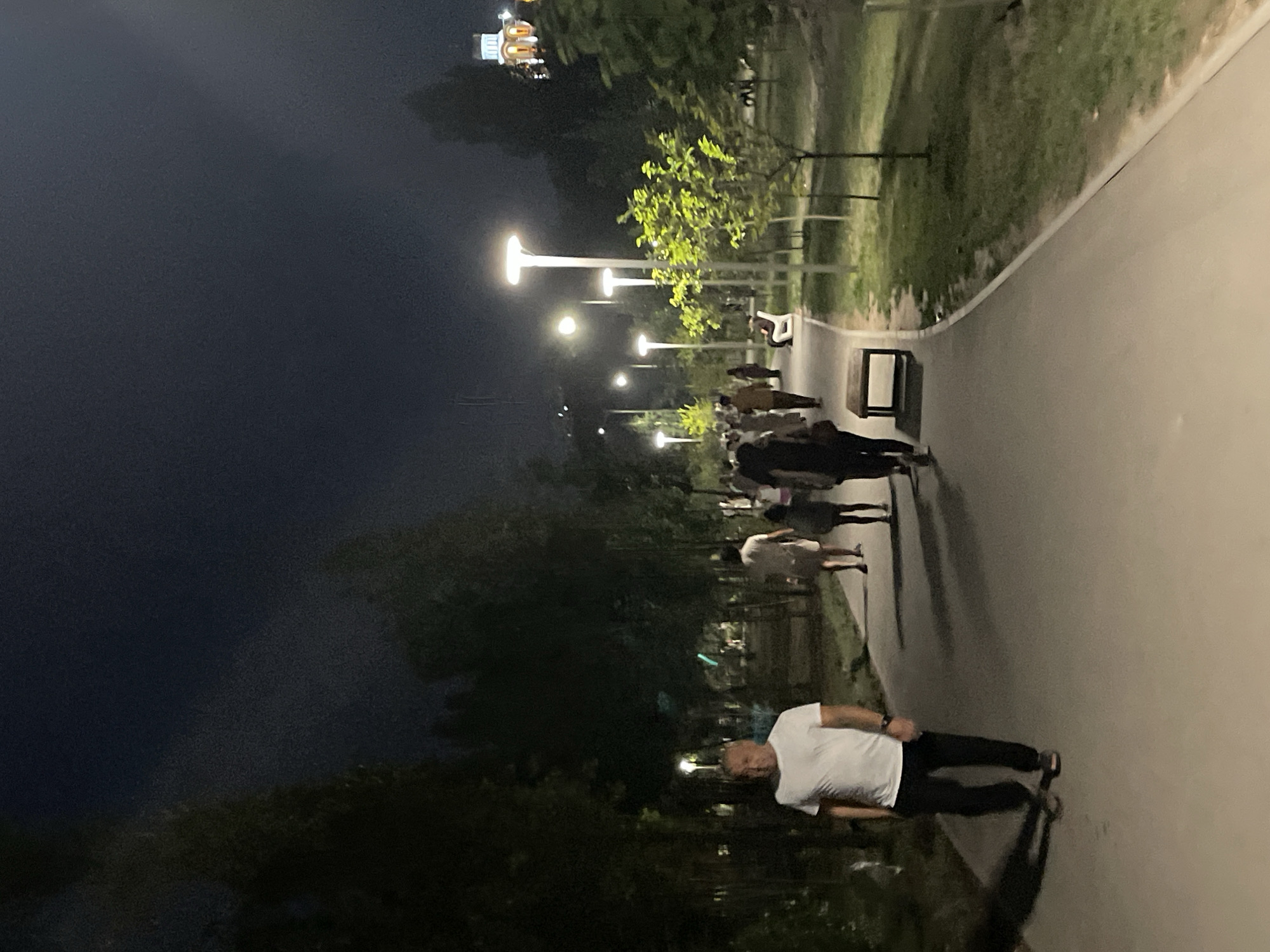 This is a nighttime photograph taken in a park in Almaty, Kazakhstan. A man in a white t-shirt and black pants is walking on a paved path. Several other people are walking in the background, some are blurred due to motion. There are lampposts illuminating the path and the surrounding trees. In the distance, a building with glowing lights is visible, suggesting it is a prominent structure within the city. The environment appears to be a public park with grass and trees.