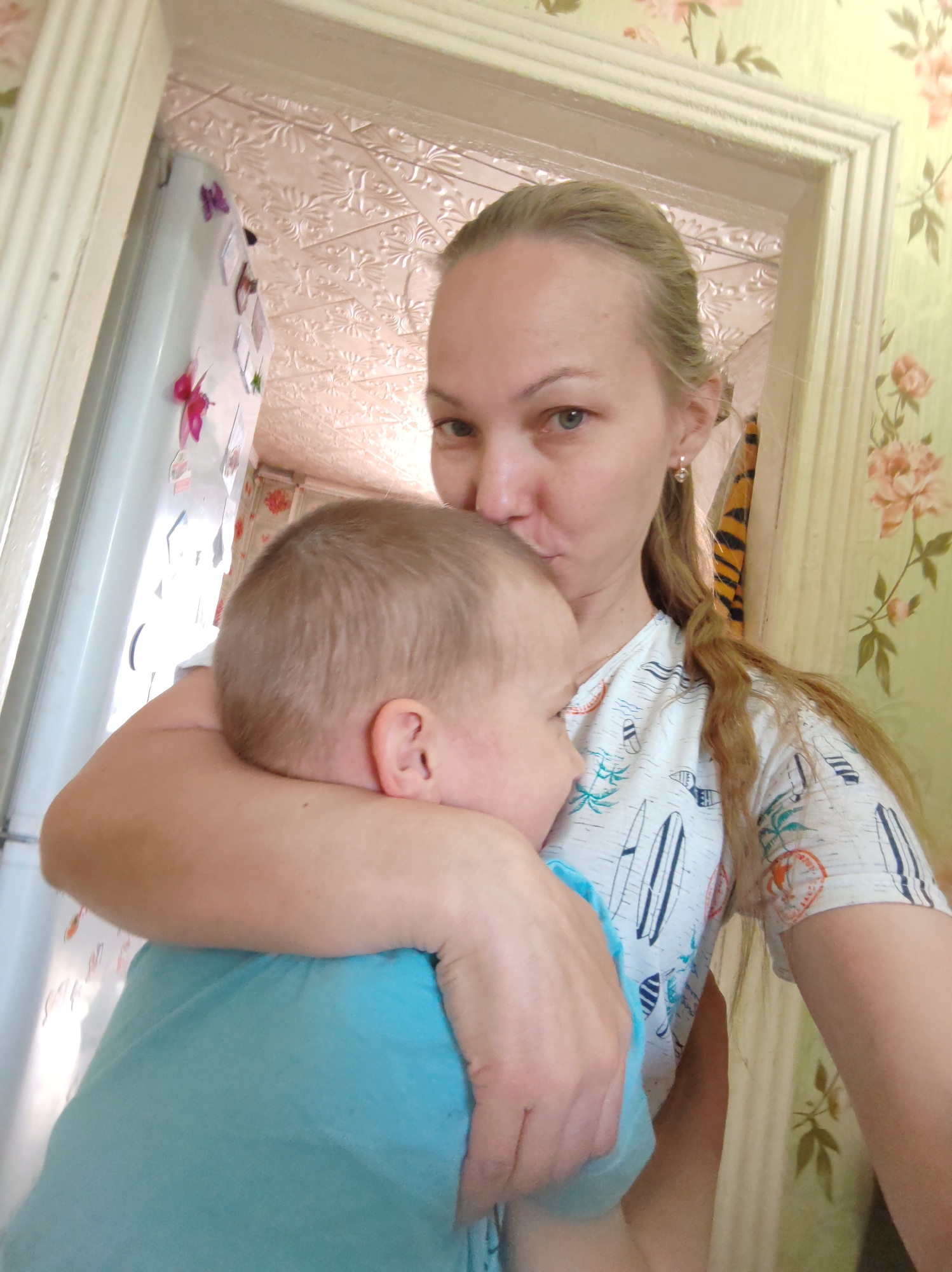 The image captures a woman indoors, holding a young child closely. She is Caucasian, with blonde hair pulled back, wearing a light-colored t-shirt with a surfboard pattern. Her expression suggests affection, as she appears to be kissing the child's head. The child, also Caucasian, has short blonde hair and is wearing a blue shirt.
The location appears to be a domestic interior, possibly a kitchen or living area. A white refrigerator with magnets is visible to the left. The walls are covered in floral patterned wallpaper. The scene is framed by a doorway with ornate molding. Based on the prompt, this scene is set in Чухлэм, Russia.