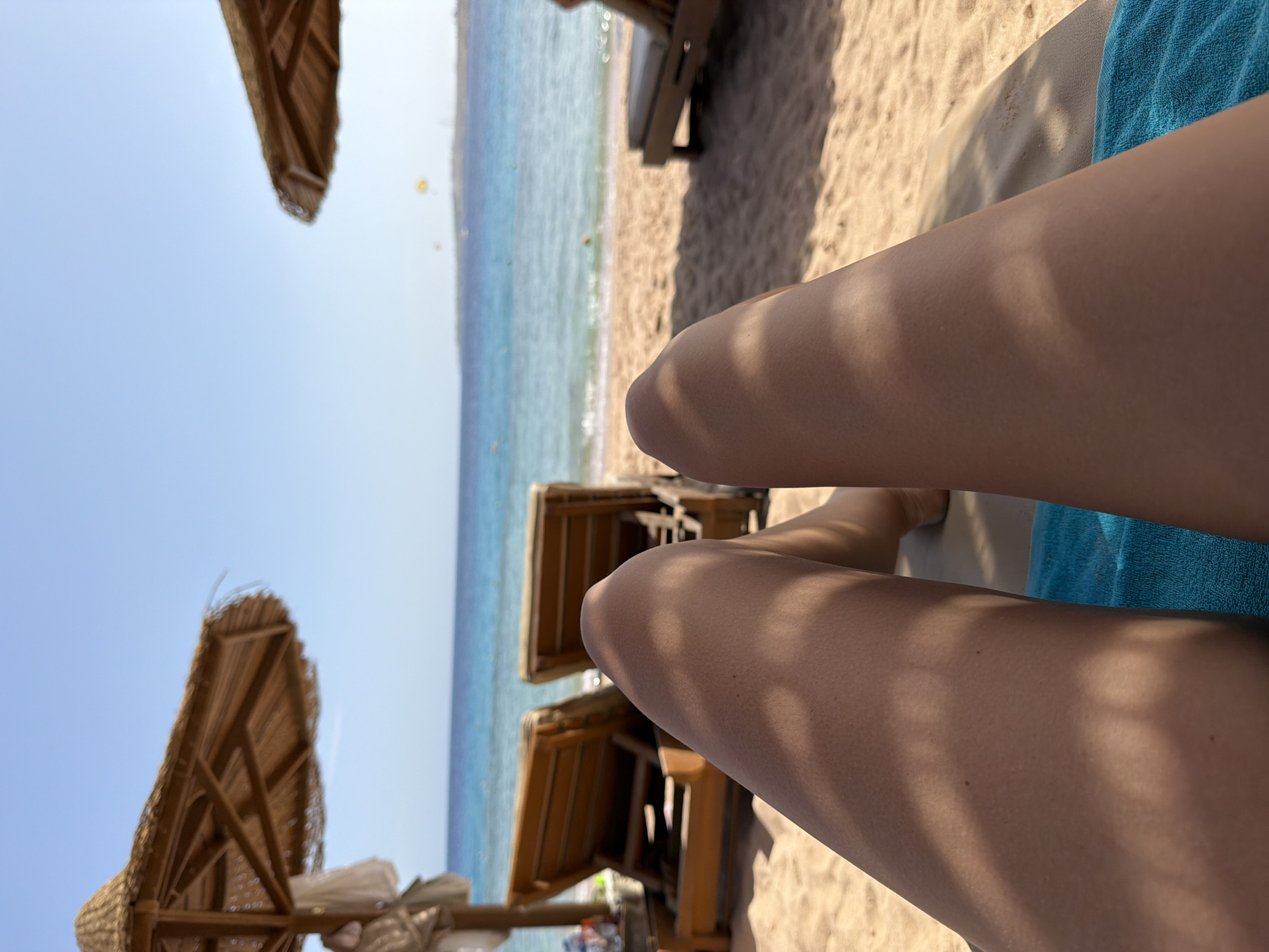 The image shows a person's legs while they are lying on a beach chair at a resort in Sharm El Sheikh, Egypt. The person is covered with a blue towel and is in the shade of a straw beach umbrella. The beach is sandy, and the sea is a clear turquoise color. Other beach chairs and umbrellas can be seen in the background. The weather appears to be sunny and warm.
