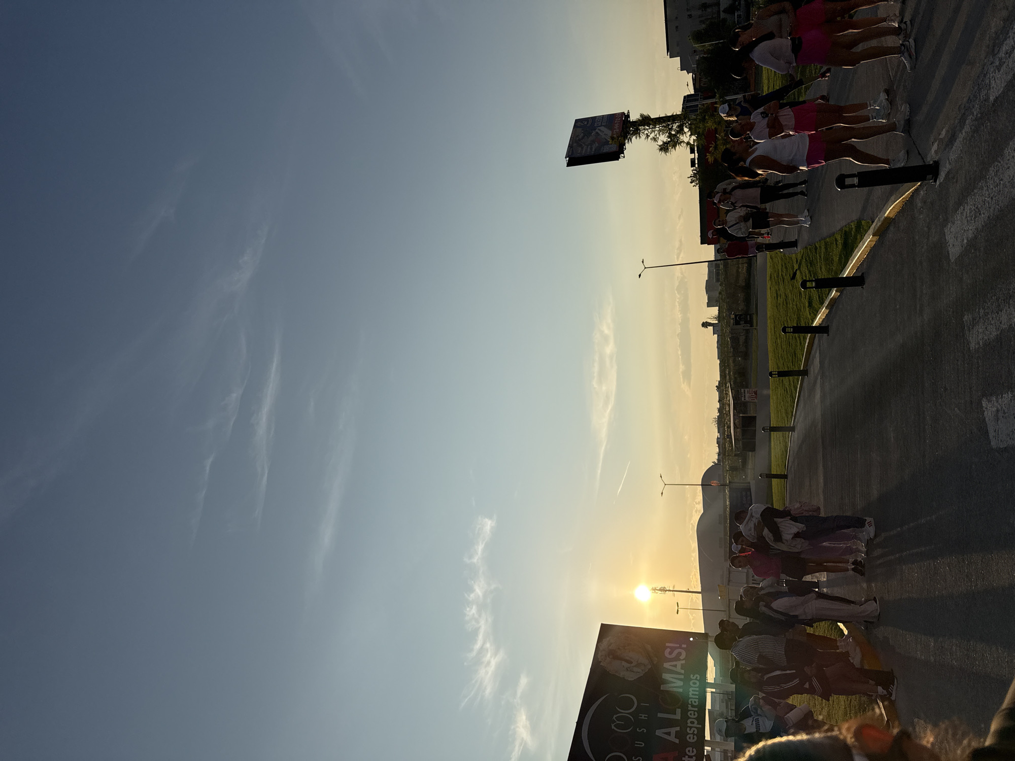 The image shows a group of people standing on a paved area beside a grassy strip, likely a sidewalk or parking lot. The group appears to be diverse, with some individuals wearing light-colored clothing and others wearing darker outfits. In the background, there's a sushi restaurant sign that says "ALOMAS!" and a large billboard. The sky is light blue and the sun is visible on the horizon, suggesting it might be early morning or late afternoon. The location is San Bernardino Tlaxcalancingo, Mexico.