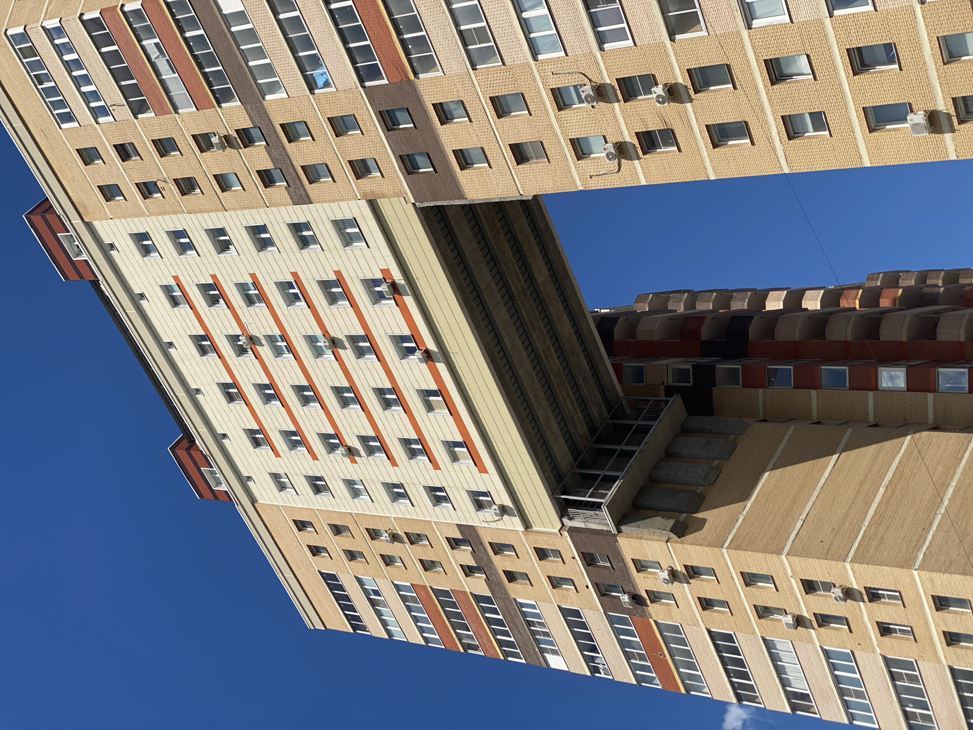 This is a photograph taken from a low angle, looking up at the exterior of a large, multi-story building against a clear, bright blue sky. The building's facade is predominantly light beige brick with alternating horizontal bands of darker brick and reddish-brown elements. Numerous windows are visible, arranged in regular patterns across the building. Some windows are open, revealing glimpses of interior spaces, while others are closed. Several air conditioning units are mounted on the exterior walls.
The building's architecture is complex and appears to be a single structure with different sections extending outwards and upwards, creating an unusual, almost cantilevered appearance in some areas. There is a distinct geometric design with sharp angles and protruding sections. A narrow, open-air staircase with stone steps can be seen ascending along one side of a protruding section of the building.
The lighting suggests it is daytime, with the sun casting shadows that highlight the building's architectural features. There are no people visible in the photograph. The overall impression is one of urban architecture and design, with a focus on the structural elements and the interplay of light and shadow on the building's surfaces. The clear blue sky indicates fair weather. The provided location context places this scene in Kotelniki, Russia. No text is visible in the image.