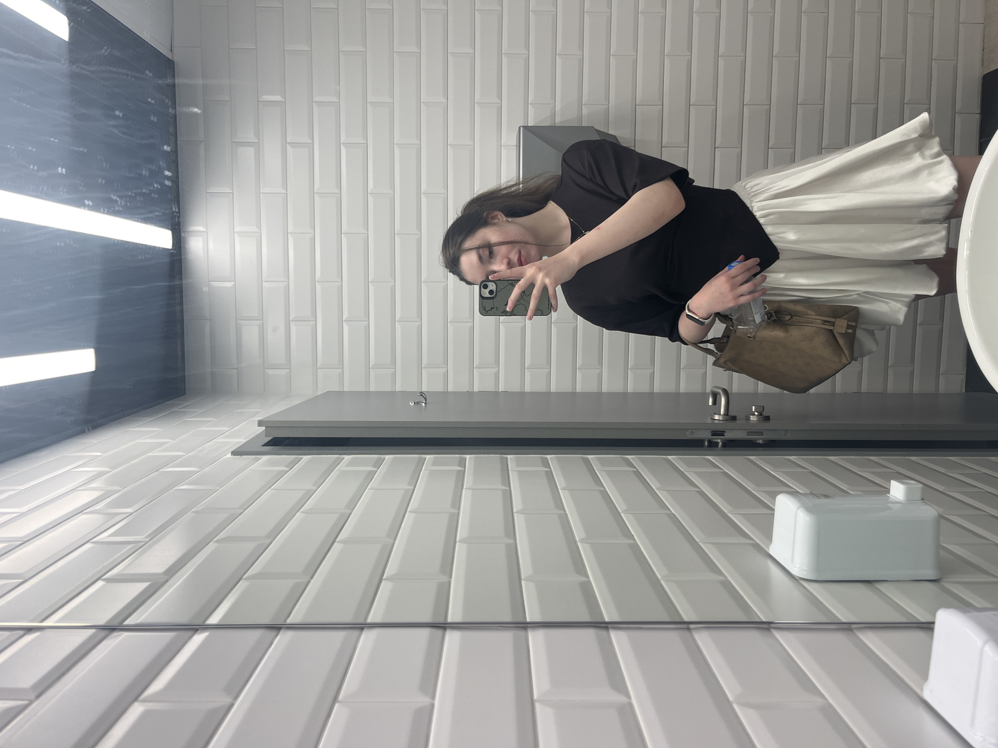A person takes a selfie in a bathroom. The bathroom is decorated with white rectangular tiles and gray countertop. There's a sink on the right. The person is wearing a black shirt and a white skirt. She's holding a brown bag. A bottle of water is partially visible. The reflection shows the mirror. The location is not apparent from the image. There is no obvious way to determine if this picture was taken in Moscow, Russia.