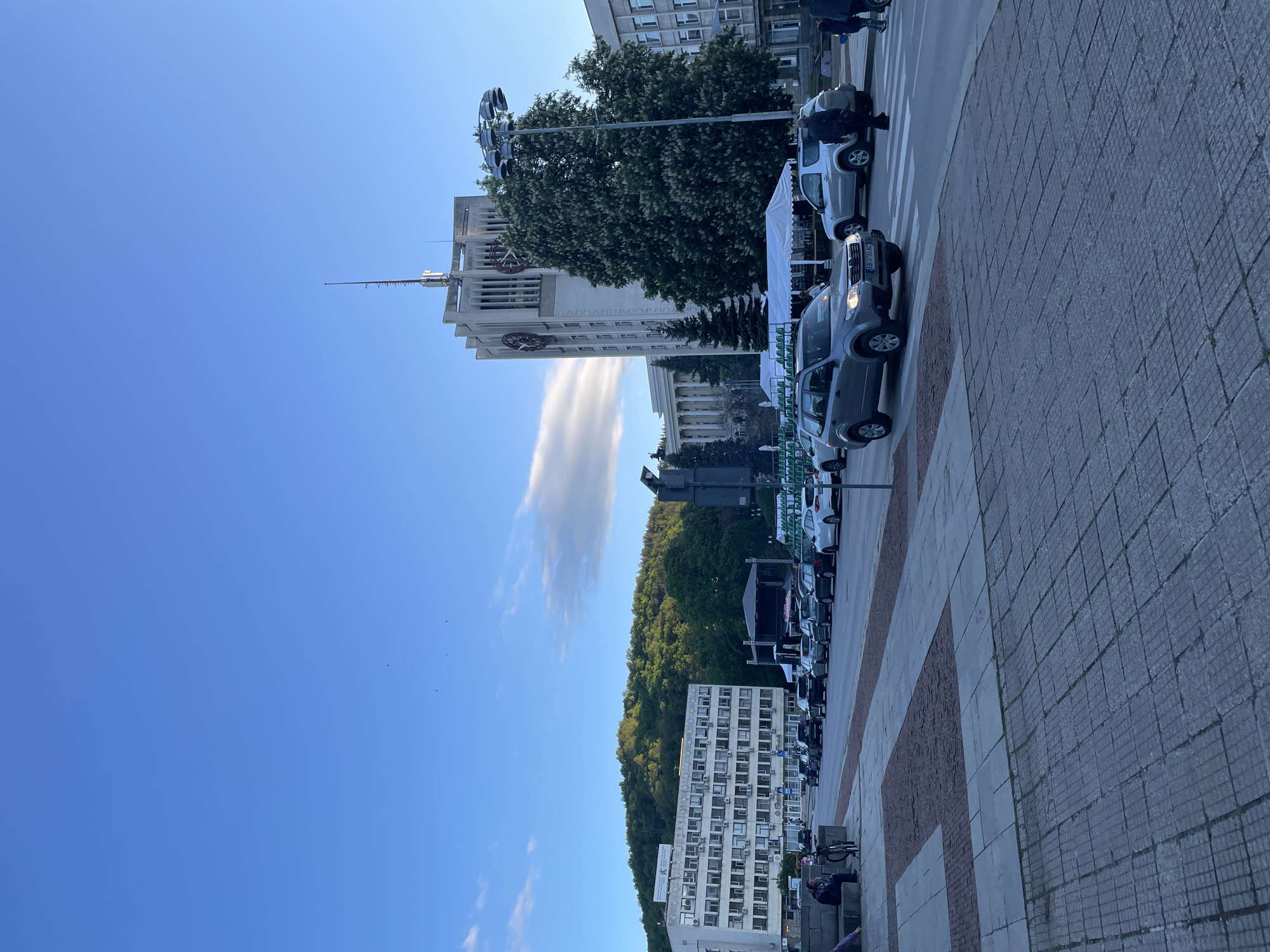 The image shows a street scene in Gabrovo, Bulgaria. A tall, light-colored building with clock faces is visible in the midground. Several cars are parked along the street, facing the same direction. On the left side of the street, there's a line of trees and a hillside covered in green vegetation. In the background, another building, multi-story and white, stands next to the forested hillside. The sky is blue with some light, elongated clouds. Pavement is in the foreground.