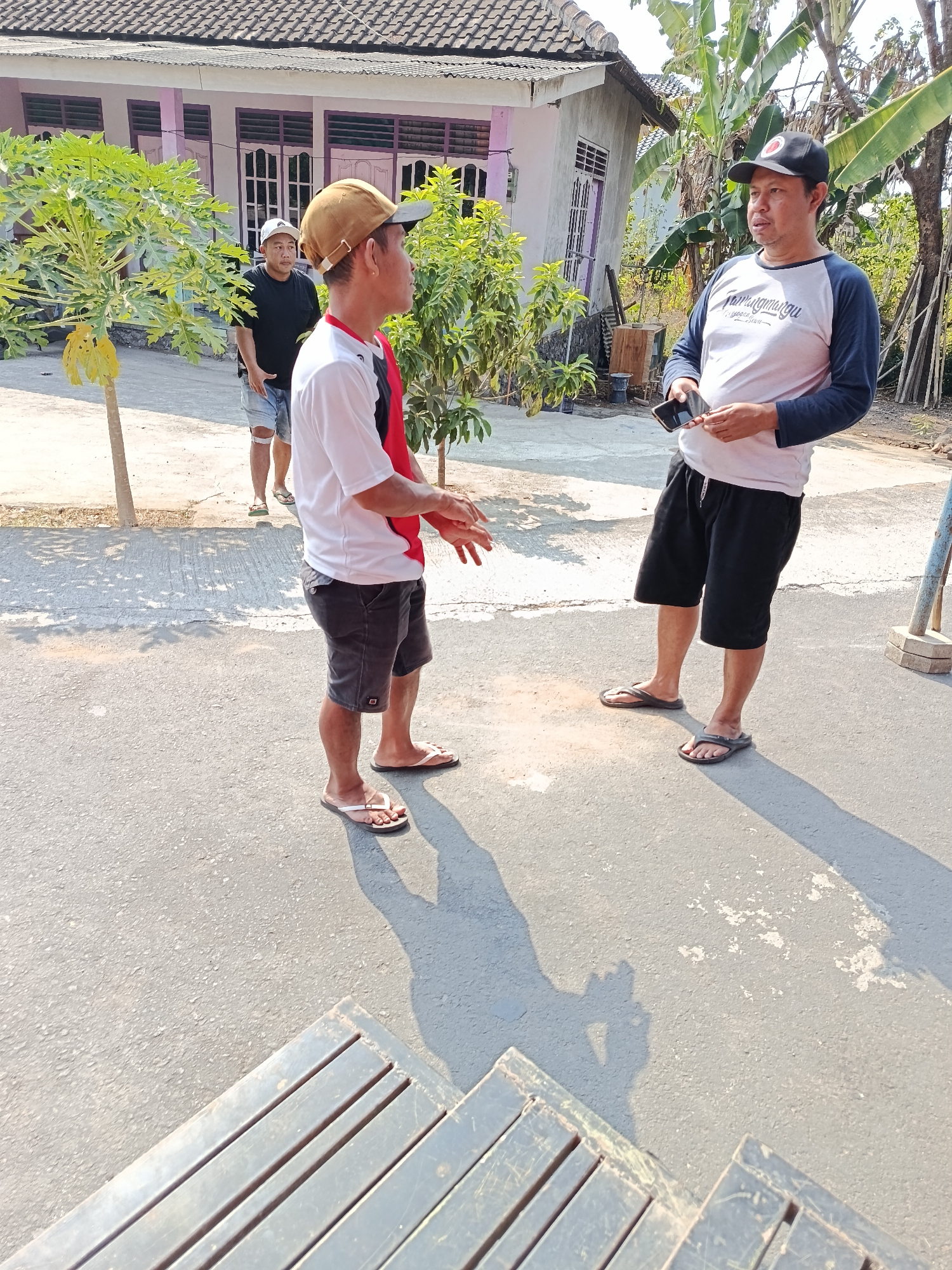 Three adult males are standing on a paved street outside a residential building in Jurug, Indonesia. The male on the left is wearing a black t-shirt, denim shorts, and a white baseball cap. He is looking towards the right, with his right hand raised in front of him. The male in the center is wearing a white t-shirt with red trim, dark shorts, and a brown baseball cap. He is facing the male on the right, with his arms extended outwards and palms up, as if speaking. The male on the right is wearing a light-colored t-shirt with blue sleeves, black shorts, and a black baseball cap. He is holding a smartphone in his left hand and looking towards the male in the center. Behind them, a two-story house with a tiled roof and purple window frames is visible, along with trees and other vegetation. The scene is illuminated by bright sunlight, casting shadows on the street.