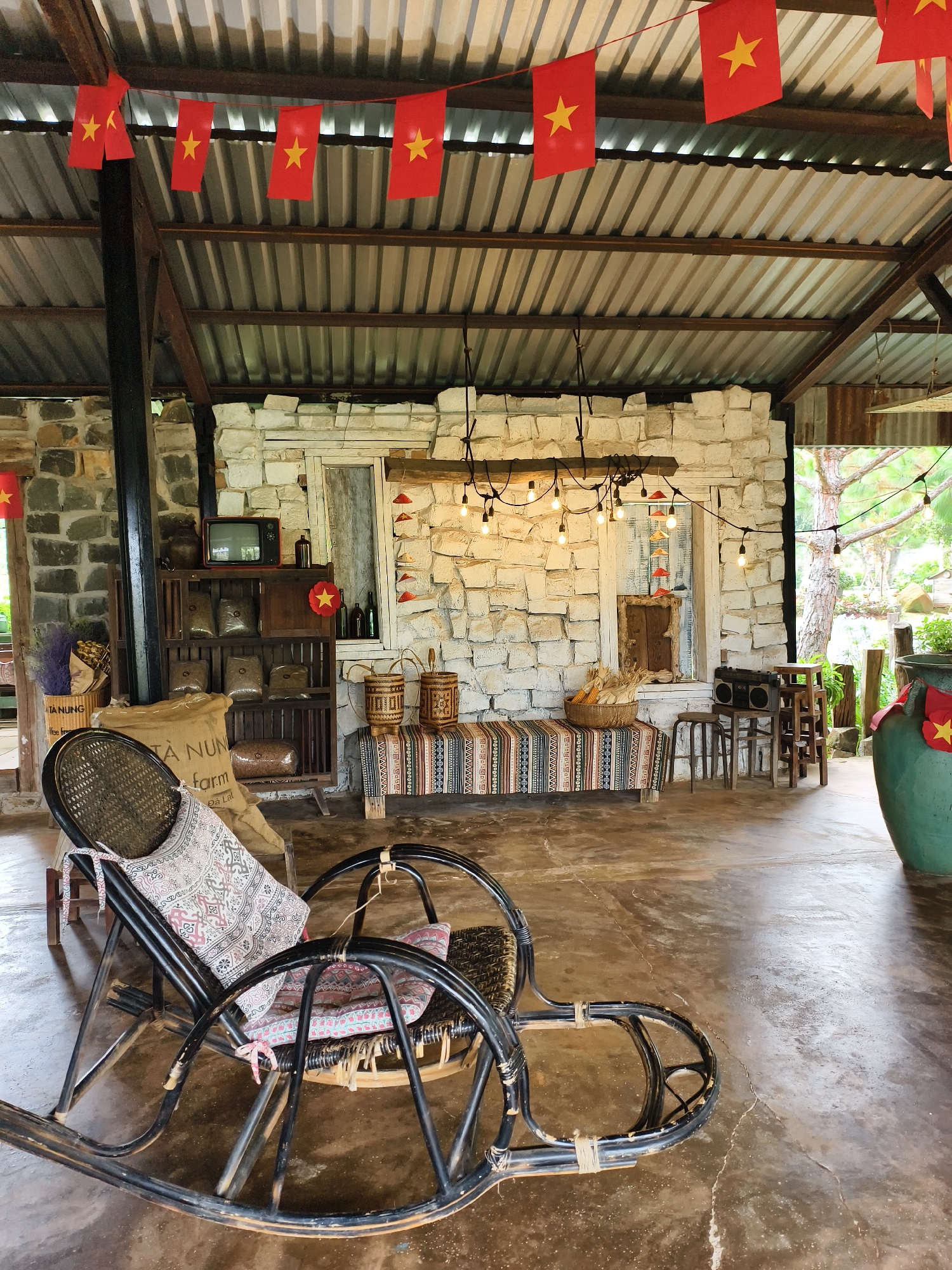 The scene is an interior space, possibly a cafe or a shop, in what seems to be Thôn 5, Vietnam. The room is characterized by a corrugated metal roof and stone walls. Red flags with yellow stars, the flag of Vietnam, are strung across the ceiling. A black rocking chair sits prominently in the foreground, draped with patterned cushions and a cloth.
Behind the chair, a wooden shelf unit displays various objects including covered bags, a vintage television, and bottles. To the right, a bench is covered with a striped textile, with baskets and corn stalks placed on top. String lights are draped above this area. Wooden stools stand adjacent to the bench. A large green ceramic pot with a Vietnamese flag is situated in the background near a window or opening. The floor appears to be concrete, with visible wear and variations in color. The environment is rustic and possibly intended to evoke a traditional Vietnamese aesthetic. There are no people visible in the scene.