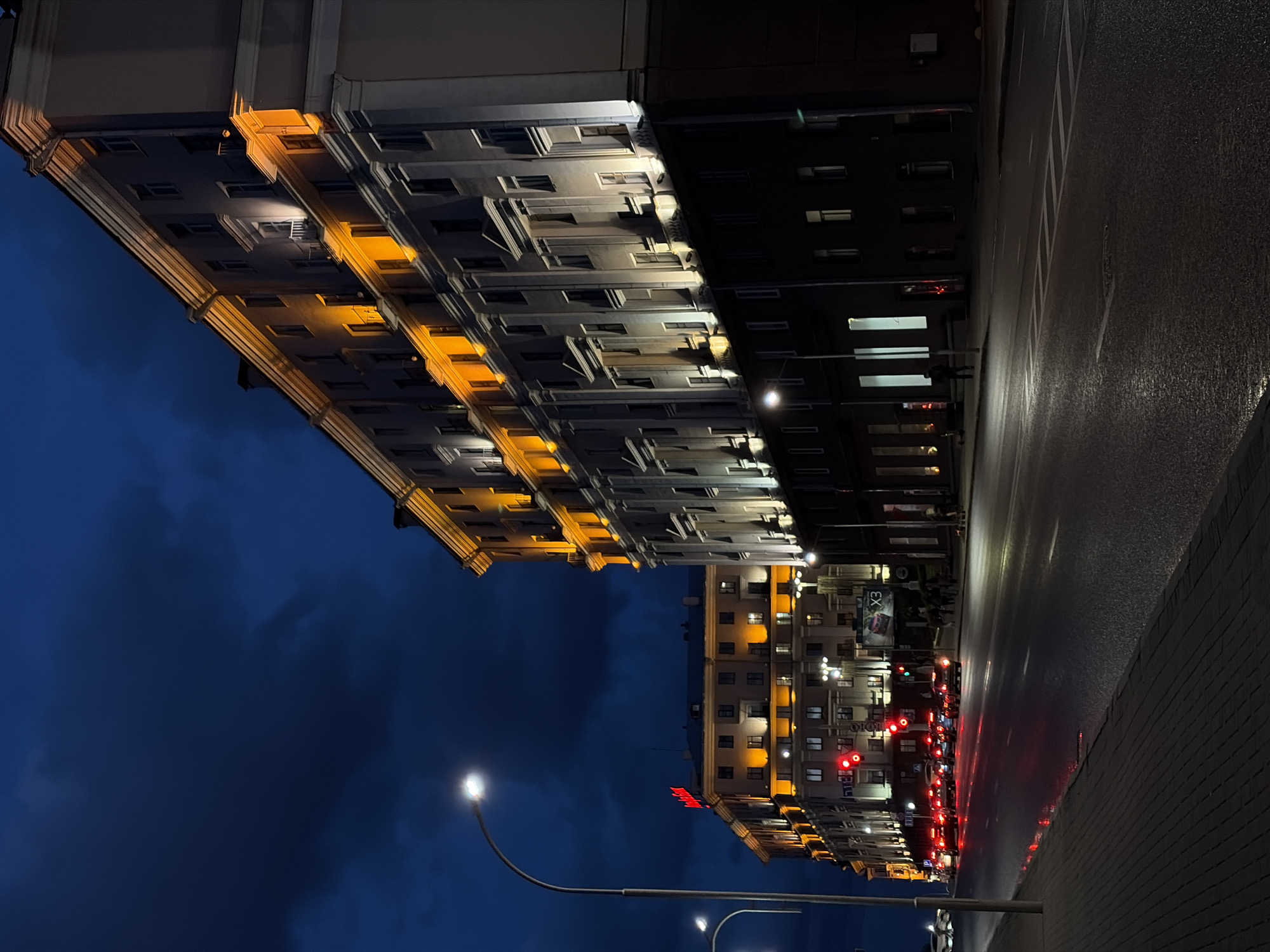 The image shows a street scene at night in Minsk, Belarus. The buildings along the street are lit up, with some having a warm, yellow light and others appearing darker. The road is wet and reflects the lights from the buildings and traffic signals. Traffic lights are visible, with several cars stopped at a red light. Street lights illuminate the scene. The sky is a deep blue, indicating it is late evening or night.