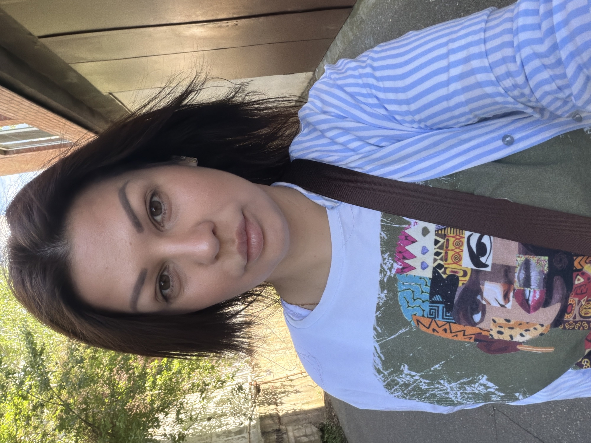 A female individual, estimated to be between 30 and 40 years old, is depicted in a selfie photograph taken outdoors in the city of Sochi, Russia. She has dark brown, shoulder-length hair, parted on the left, which appears slightly wind-blown. Her facial features include dark brown eyes, dark and groomed eyebrows, a straight nose, and full, naturally colored lips that are slightly parted. She is wearing a white short-sleeved t-shirt featuring a large, multi-colored graphic print of a stylized, abstract face composed of various geometric and textured patterns. Over this t-shirt, she wears an unbuttoned, light blue and white striped long-sleeved shirt with the sleeves rolled up to the elbows. A dark brown strap, consistent with a shoulder bag, crosses her torso diagonally from her right shoulder. A thin, light-colored chain is visible around her neck, beneath the collar of her t-shirt. A small, light-colored earring is visible on her right ear.
The background to the viewer's left consists of a concrete wall, likely part of an underpass or retaining structure, with sunlight casting distinct shadows of tree foliage upon it. Green vegetation is visible above and behind this wall. The background to the viewer's right features a structure with dark brown vertical panels, and above it, a section of a brick building with a window is visible. The ground appears to be a paved or asphalt surface. The lighting indicates direct sunlight, suggesting either morning or late afternoon.