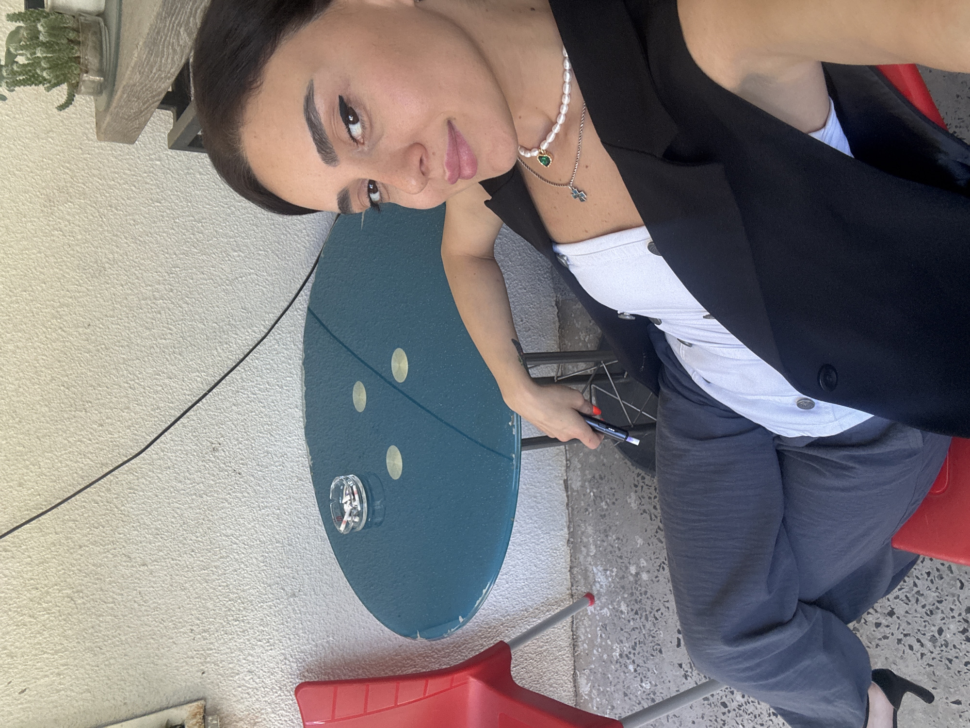 The image depicts a woman sitting outdoors at a small, circular, teal-colored table. An ashtray filled with cigarette butts sits on the table. To the left of the table is a red chair with a metal frame. The woman is wearing a white top and a black vest, with grey pants. She has on dark-rimmed glasses and a pearl necklace with a green pendant and a cross charm. She is holding a dark-colored pen or stylus in her right hand, with bright red nail polish visible on her fingernails. The background shows a textured, off-white wall, and a plant in a pot on a wooden shelf is visible to the upper right. The ground is a speckled concrete surface. The scene appears to be set on a patio or outdoor seating area.