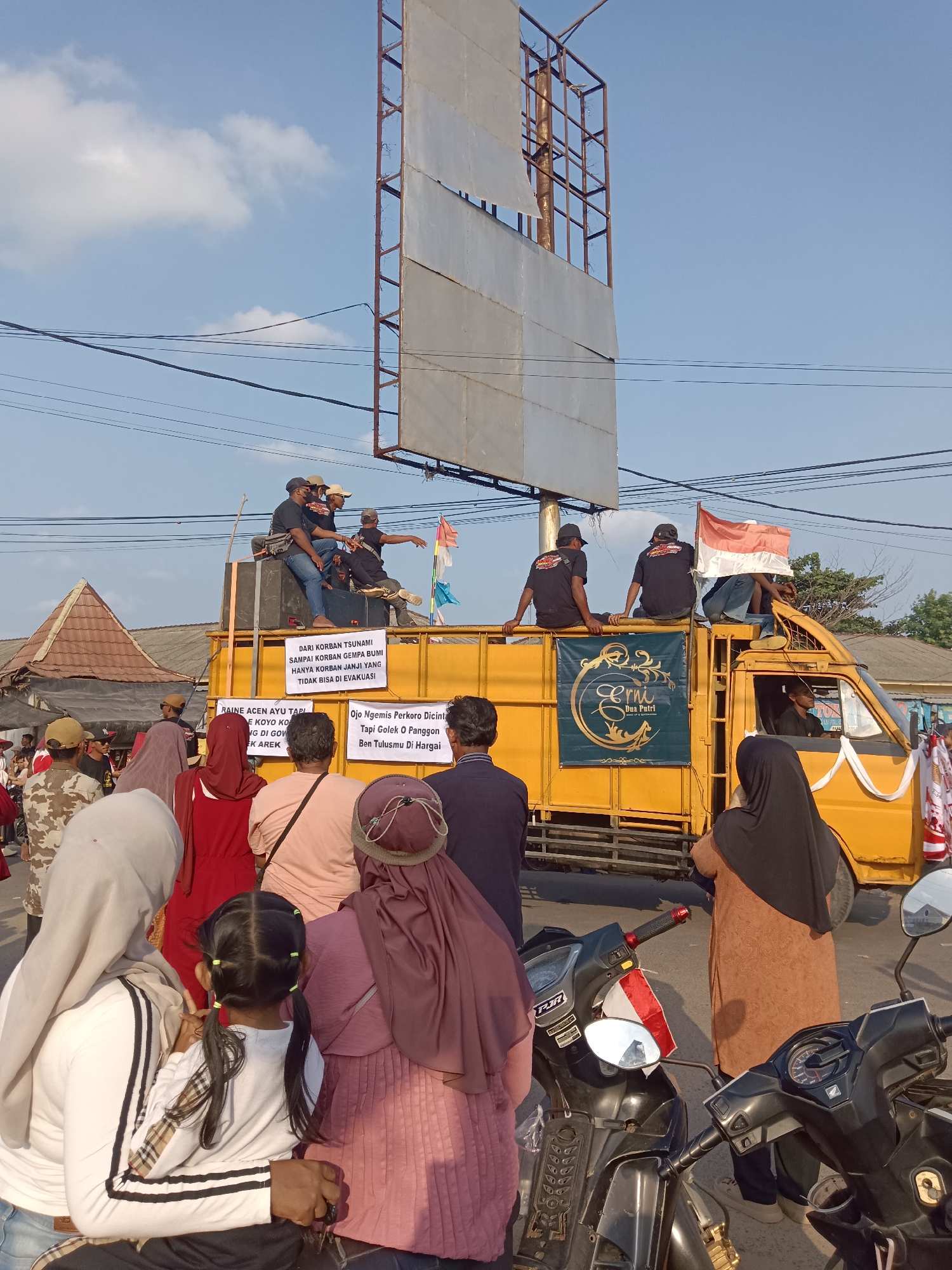 A group of people are gathered on and around a yellow truck in Tegal Gondo, Indonesia. The truck has several signs displayed on it. One sign reads "DARI KORBAN TSUNAMI SAMPAI KORBAN GEMPA BUMI HANYA KORBAN JANJI YANG TIDAK BISA DI EVAKUASI". Another sign on the truck has the text "Erni Dua Putri" in gold lettering on a dark green background. A group of men are sitting on top of the truck, some wearing black t-shirts. An Indonesian flag is visible on the truck. In the foreground, several individuals are standing, some wearing headscarves. Two motorcycles are parked in front of the truck. The sky is blue with scattered clouds. A billboard structure is visible behind the truck.