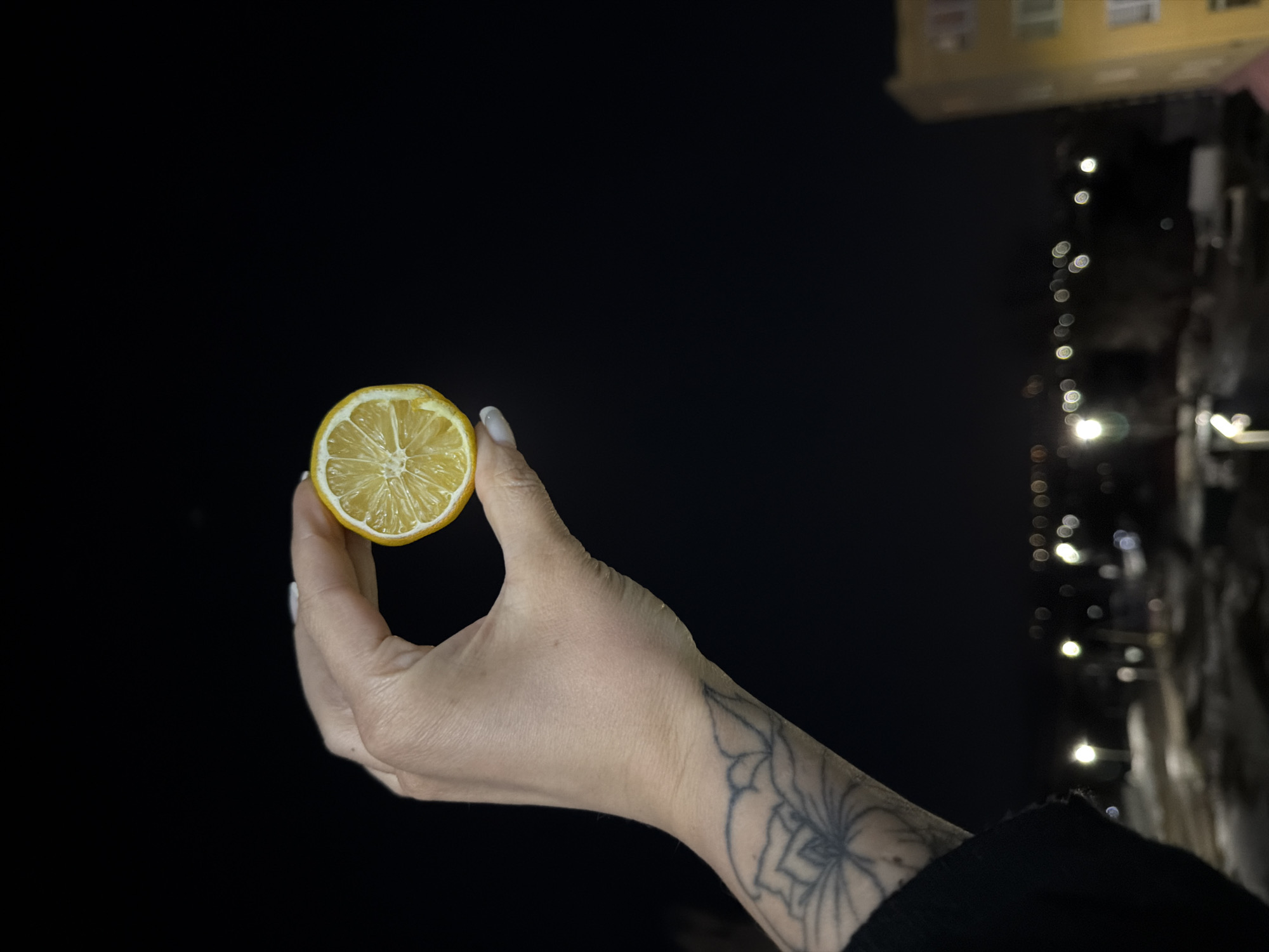 A human hand, adorned with a tattoo, holds a half-cut lemon. The background is a night scene with a blurred building and street lights. The location is most likely in Sarapul, Russia.