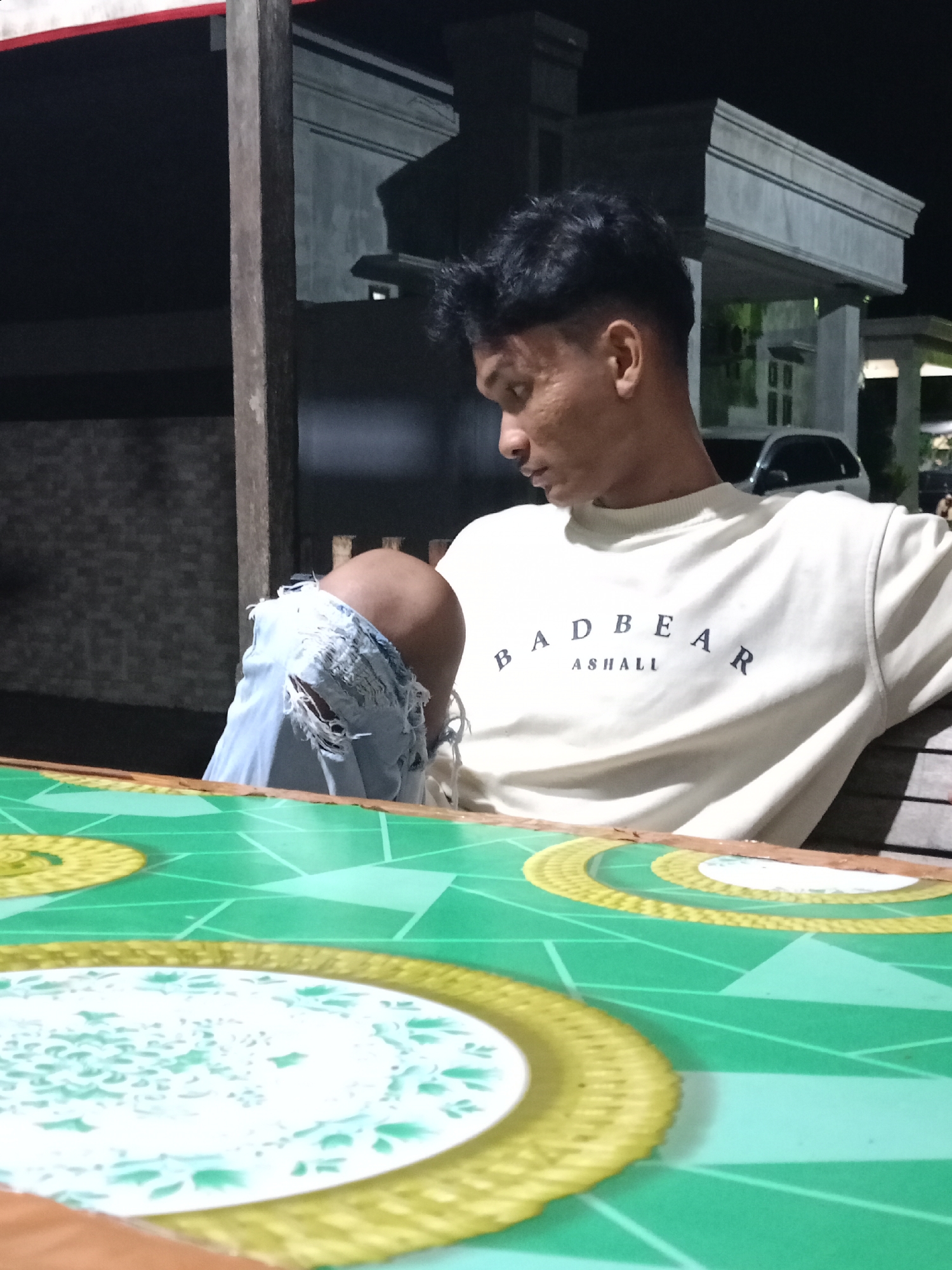 A young Asian man sits outdoors at night in Lhokseumawe, Indonesia. He is wearing a cream-colored "BAD BEAR ASHALL" sweatshirt and ripped light blue jeans. He is seated at a table that has a bright green surface with geometric patterns and circular, decorative elements in gold and white. The man is looking down and to his left. In the background, there is a building with a lit porch and a car parked nearby. The area appears to be dimly lit. A wooden post is visible near the left edge of the frame.