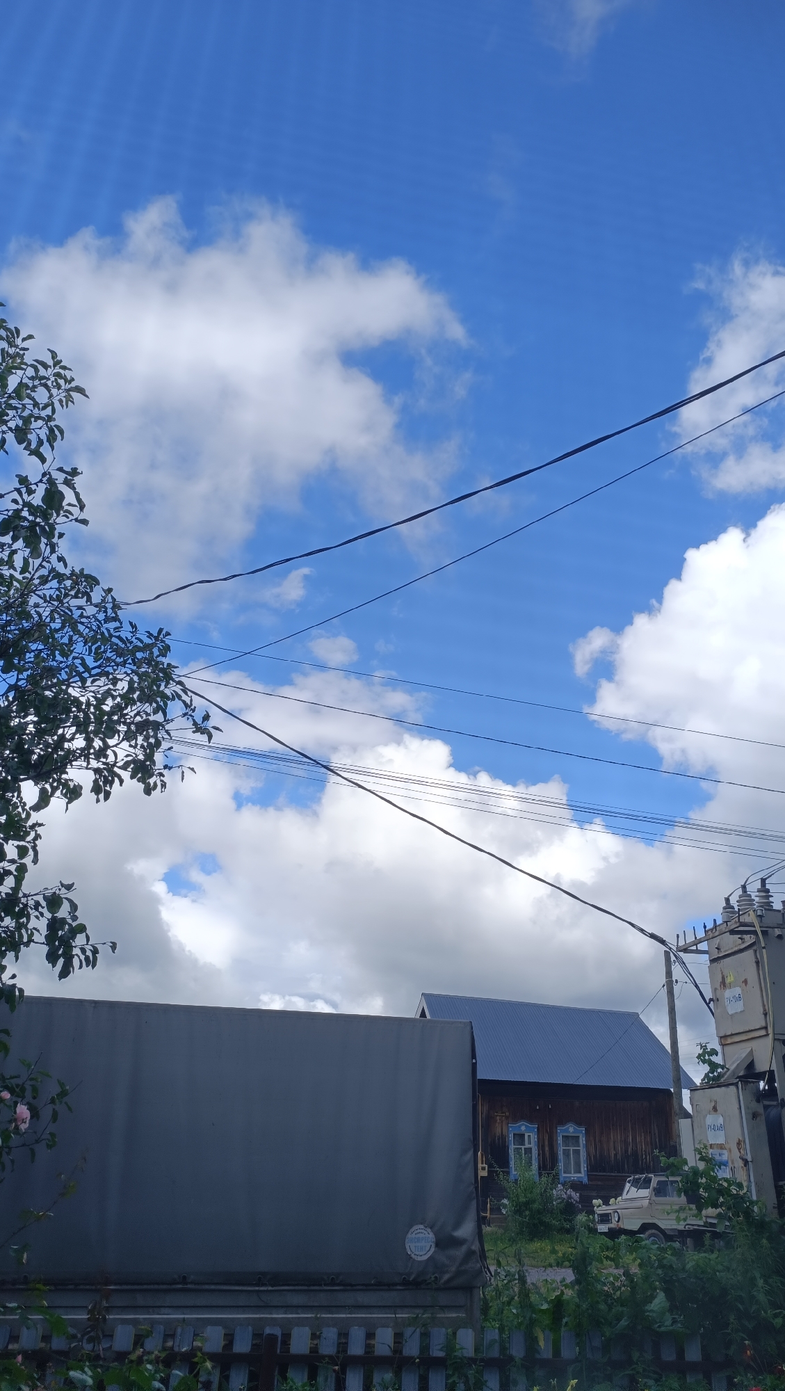 The image depicts a rural scene with a bright blue sky dotted with white, fluffy clouds. In the foreground, the top of a large, gray tarpaulin-covered truck is visible, along with a portion of a weathered wooden fence. Behind the truck, a traditional wooden house with a blue metal roof stands, featuring decorative blue window frames. To the right of the house, a small, light-colored off-road vehicle is parked. A utility pole with electrical equipment and wires is also present in the background. Foliage from trees and bushes surrounds the structures. The overall impression is one of a sunny day in a village setting, possibly in the city of Karsha, Russia, given the architectural style and the presence of power lines and a transformer.