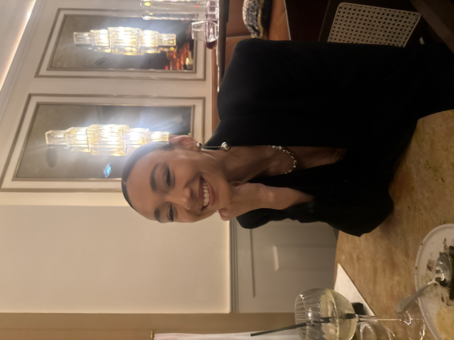 A female subject, appearing to be in her 20s or 30s, is seated at a table in an indoor restaurant identified as Le Relais Plaza, located within the Hôtel Plaza Athénée at 25 Avenue Montaigne, 75008 Paris, France. She is positioned slightly right of center, looking directly at the camera with a broad smile. Her right hand is propped under her chin. She wears her dark hair pulled back, a black blazer, a silver-toned chunky necklace, and a long, dangling earring with two spherical elements on her left ear. On the light-brown table in the foreground, to the left, is a stemmed glass containing a light-colored beverage with a black straw. Immediately in front of the glass, a small white plate with dark food remnants and a silver spoon is visible. The background features light-colored walls with paneling on the left. Two large, framed mirror artworks are centrally positioned, each containing an elongated, tiered crystal light fixture that reflects bright light. A dark, upholstered banquette is partially visible on the far right. The overall lighting is warm and ambient.
