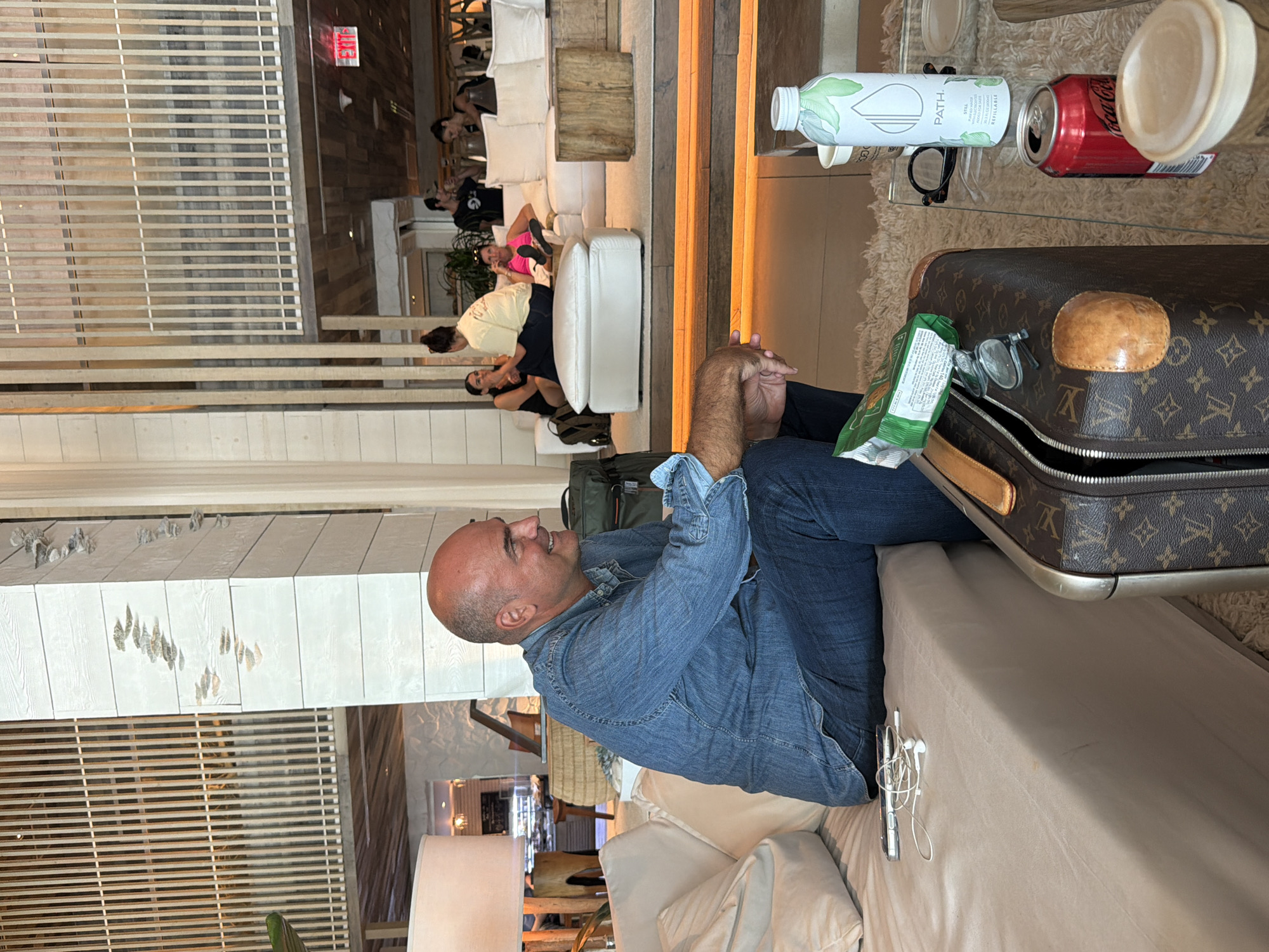 This image captures an indoor lounge or waiting area, likely within a hotel or similar establishment, in Miami Beach, USA. The lighting is warm and ambient, suggesting evening or a deliberately relaxed atmosphere.
In the foreground, a bald man wearing a denim shirt and jeans is seated comfortably on a light-colored sofa, facing to his right with a slight smile. His hands are clasped. Next to him, on the sofa, is a vintage Louis Vuitton monogrammed suitcase, partially unzipped. Inside or near the suitcase are a green snack bag with illegible text, a pair of eyeglasses, and a smartphone with white wired earbuds.
On a clear glass table in front of the man, a white "PATH." brand water bottle with green leaf designs is visible, bearing the text "REFILLABLE PURIFIED WATER." Beside it is an open red "Coca-Cola" can and another pair of eyeglasses.
The background reveals several other individuals seated on modern white modular sofas and chairs, some appearing to be conversing. One woman is distinct in a bright pink top. The decor features light-colored wooden slatted walls, wooden steps or platforms, and subtle indirect lighting. A red "EXIT" sign is visible on a wall in the distance. The scene depicts a casual yet upscale social or waiting environment.