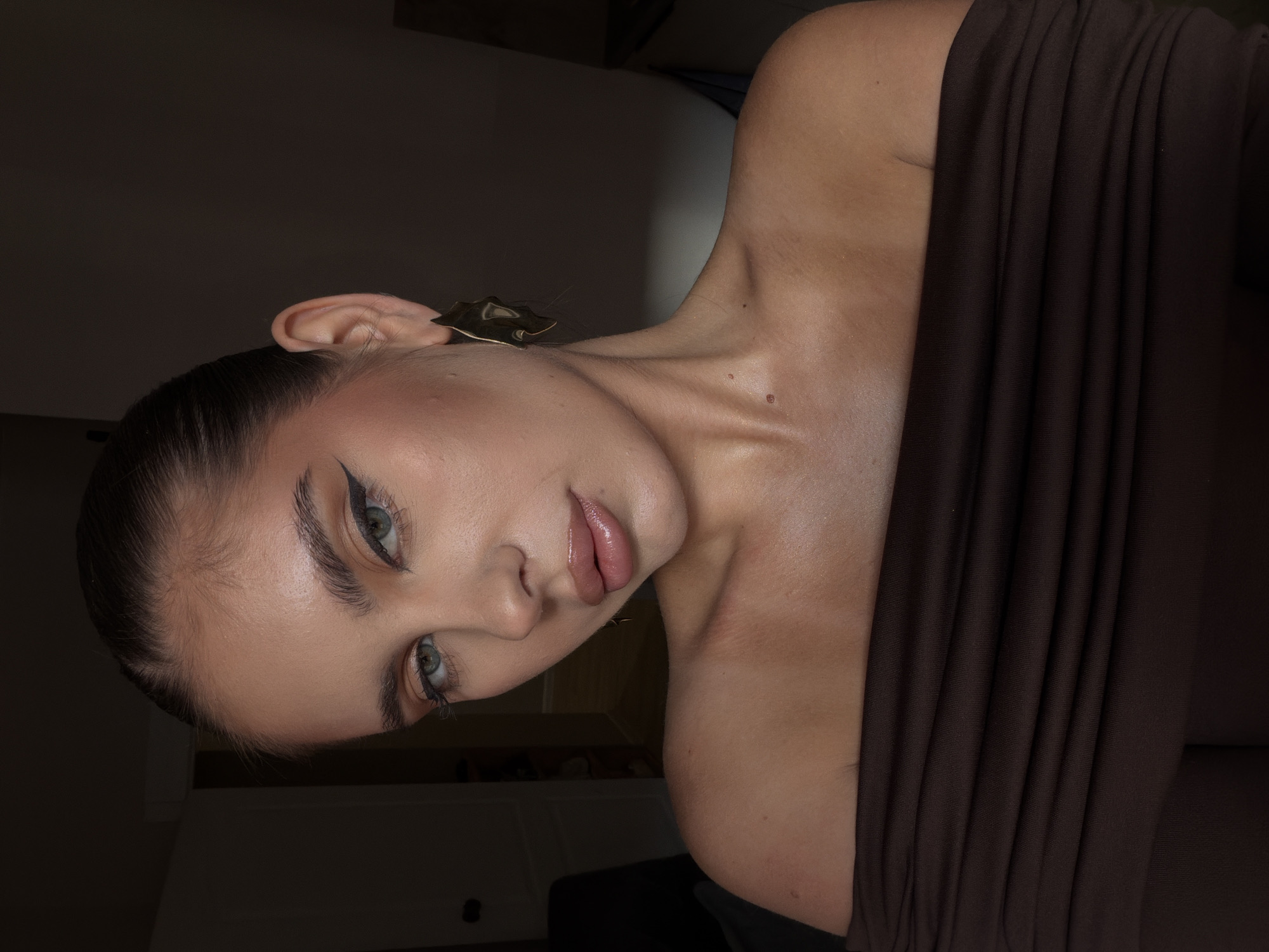 A young woman is depicted in a portrait. The subject's features include blue-green eyes, defined eyebrows, and a slicked-back hairstyle. Her skin has a noticeable sheen. She wears a dark brown off-the-shoulder top. A gold leaf-shaped earring is visible on her left ear. The background is dimly lit with a light source highlighting the subject. The image does not include the location or any information that leads to the city of Krasnoyarsk, Russia.