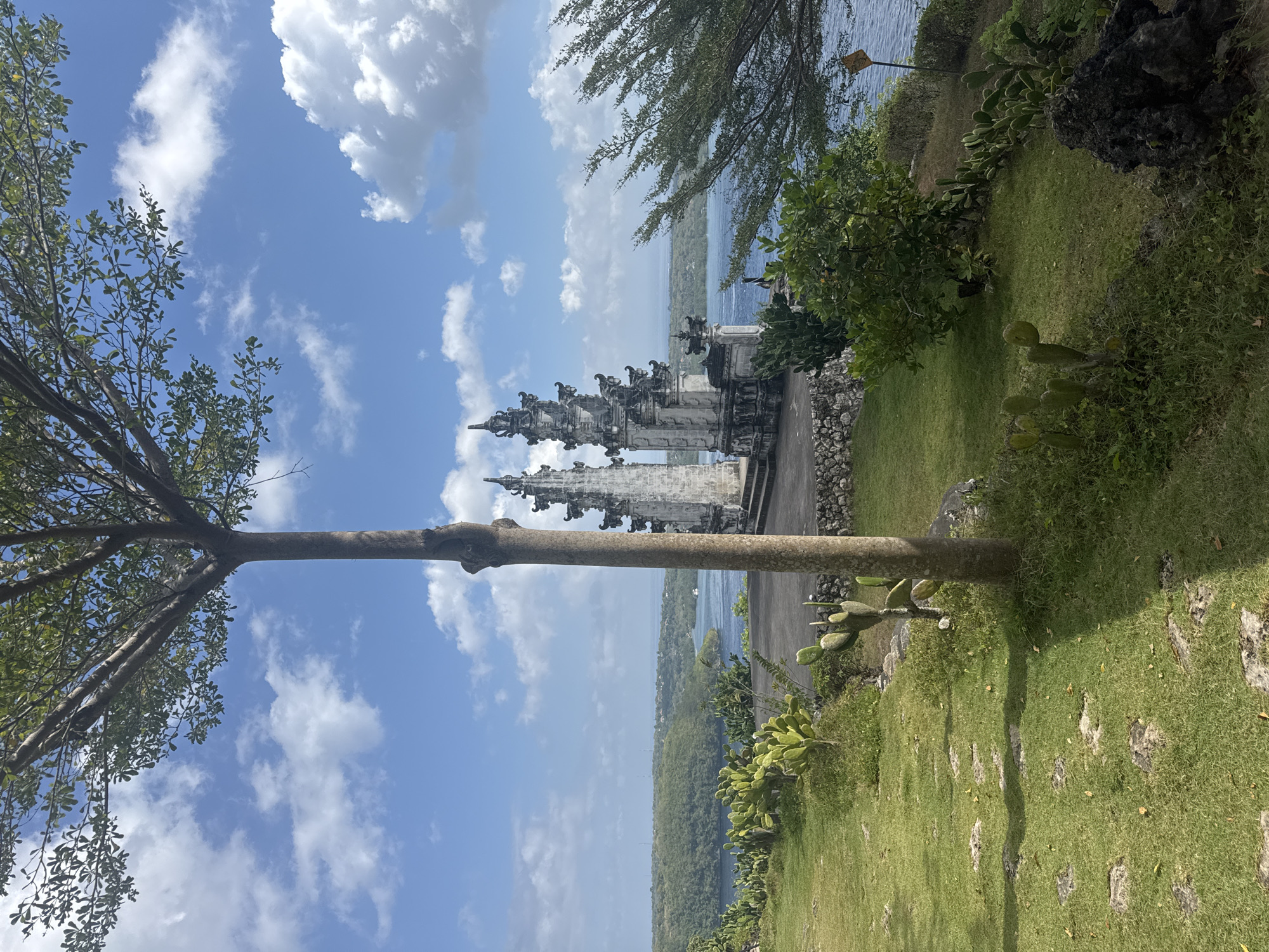 A bright, clear day illuminates a scenic view of the ocean and coastline, with a Balinese temple structure prominently featured. The temple, built of light-colored stone, has multiple tiered roofs and ornate carvings. It is situated on a grassy hill overlooking a wide expanse of blue water, which meets a lush, green, and hilly landscape in the distance. The foreground is a manicured lawn dotted with cacti and a single, slender tree in the center. The tree's trunk is thick and its branches reach upwards, partially obscuring the bright blue sky filled with white, puffy clouds. The ocean is calm and reflects the sky. A small boat is visible on the water. The overall scene conveys a sense of tranquility and natural beauty. The location is the Thousand Islands Viewpoint (Pulau Seribu) in Nusa Penida, Indonesia.