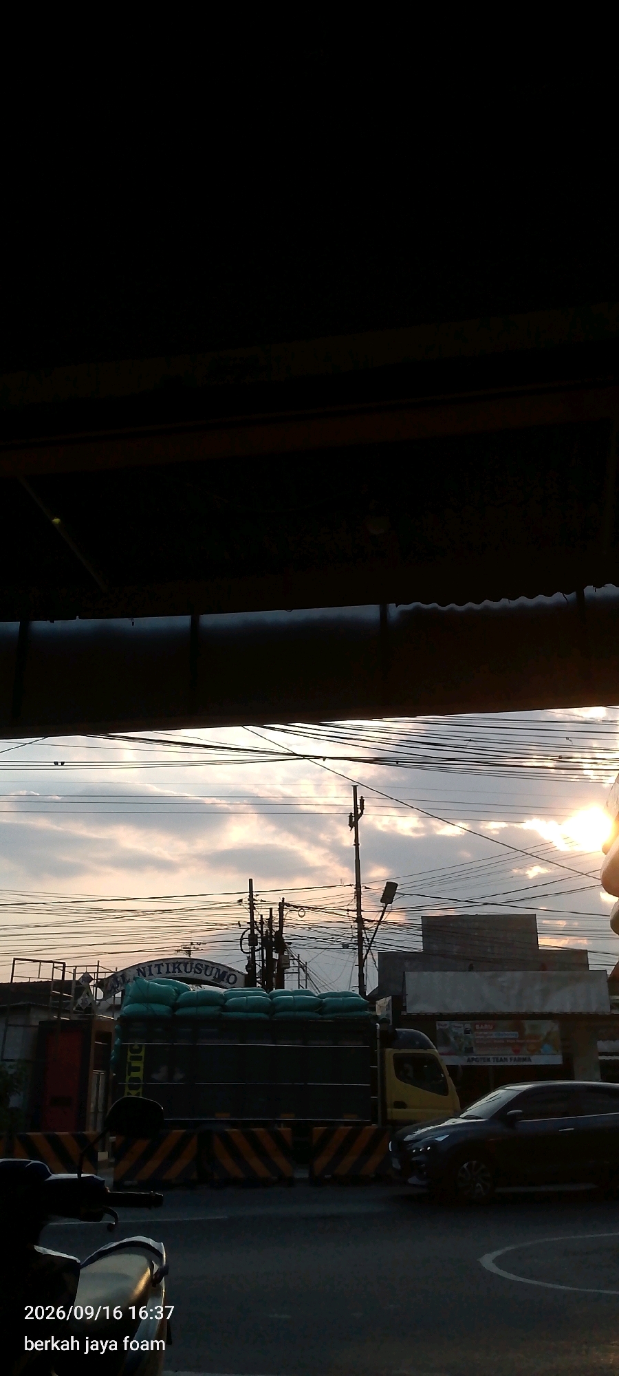 The image captures a street scene in Madiun, Indonesia, during what appears to be late afternoon or early evening, given the low angle of the sun and the lighting conditions. The foreground is dominated by the front of a motorcycle, partially obscuring the view. The timestamp "2026/09/16 16:37" and the text "berkah jaya foam" are overlaid in the bottom left corner, suggesting this is a personal photograph or a timestamped record.
The midground features a road with a black sedan in motion on the right side. To the left of the sedan, a large truck is parked or moving slowly. The truck is laden with numerous large, light green sacks, likely containing goods. Behind the truck, a sign with the partially visible text "AL NITIKUSUMO" can be discerned. The truck appears to be bordered by a yellow and black striped barrier.
The background reveals a sky with scattered clouds illuminated by the setting sun, creating a soft, diffused light. Several utility poles with a dense network of electrical wires crisscross the sky, a common sight in urban Indonesian environments. Various buildings, some appearing to be shops or commercial establishments, are visible in the distance. The overall impression is of a busy urban setting at the end of the day.