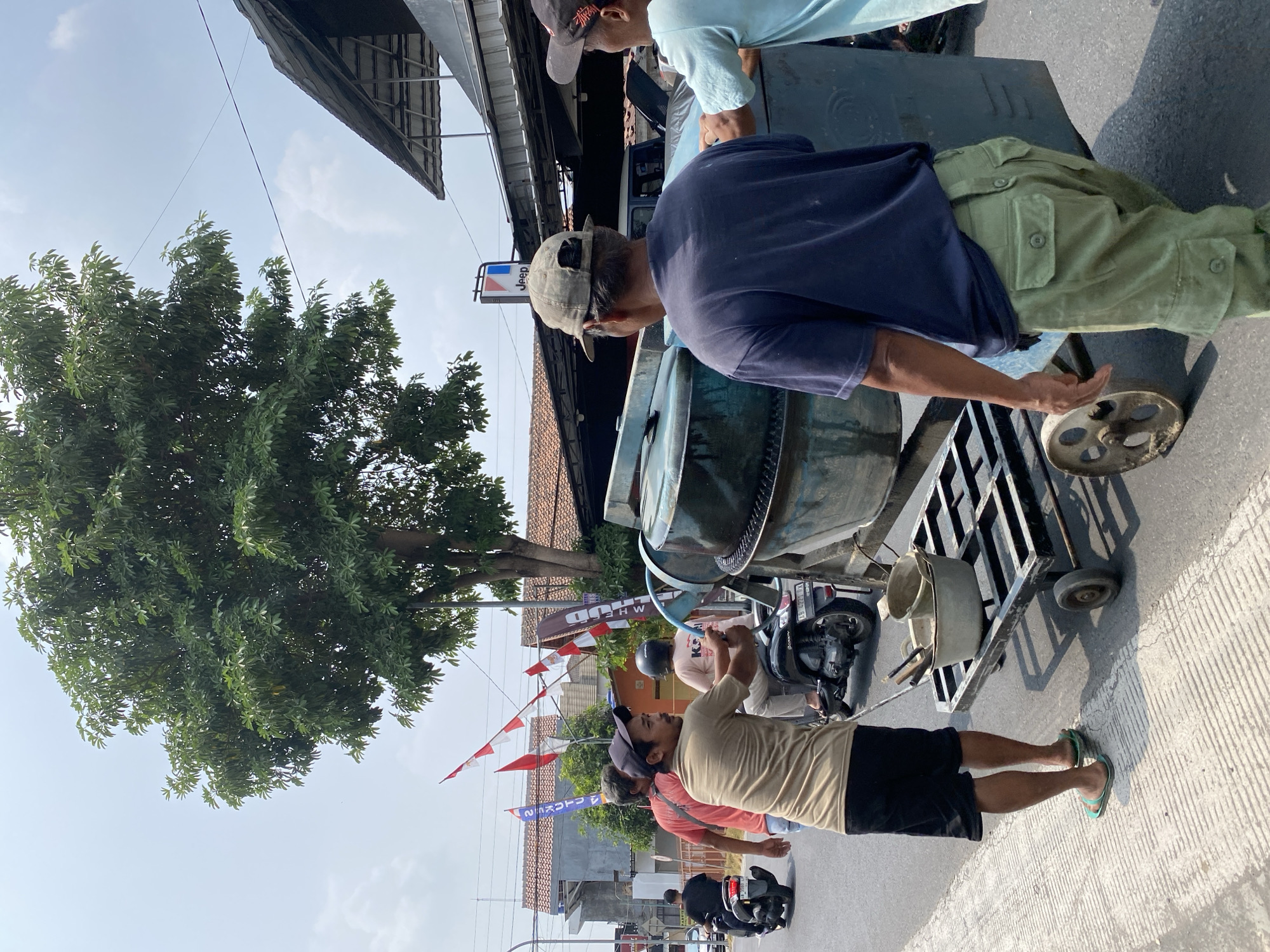 In Kasihan, Indonesia, on a sunlit day, a group of men are working on a street. The scene is set along a road with a concrete curb and asphalt pavement. In the foreground, a man wearing a beige t-shirt and black shorts, with green flip-flops, pushes a metal cart that carries two buckets, one containing construction tools. Behind him, a large, blue cement mixer, likely being moved, is a central object. Two men are interacting with the cement mixer, one in a dark blue t-shirt and light green cargo pants, and another in a light blue t-shirt.
In the background, a variety of elements indicate a bustling environment. Indonesian flags, red and white, are seen flying from poles. Several buildings with terracotta roofs are visible, suggesting a residential or commercial area. A Jeep dealership sign is present, with the brand's logo displayed. Motorcycles are on the road, with one man on a black scooter in the left midground. Another scooter with a rider is near the cement mixer. A large tree with dense green foliage dominates the upper left portion of the image. The sky is bright and partly cloudy.