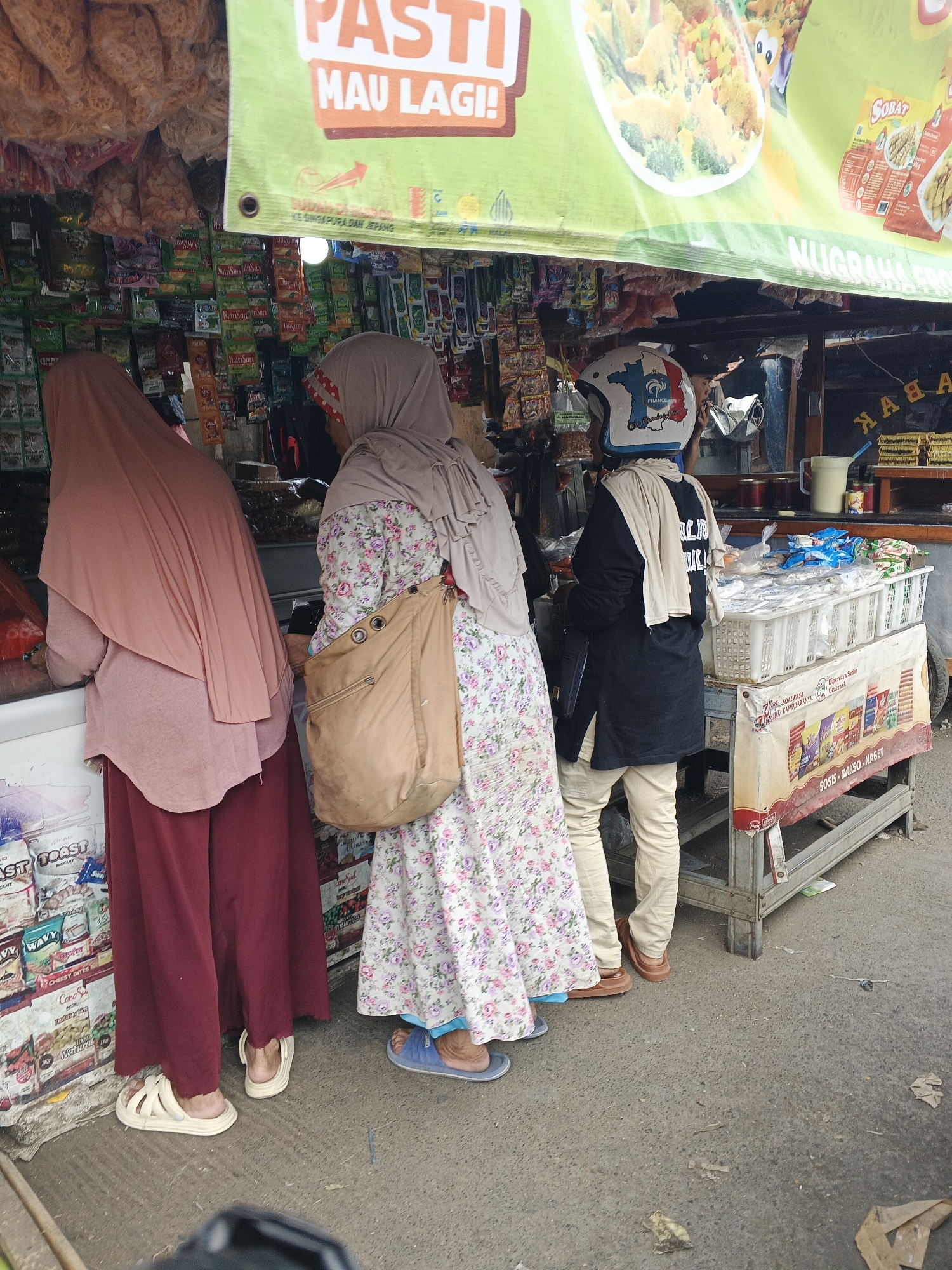 The image shows a street-level view of a small food stall in Sukabumi, Indonesia. Three individuals are present. Two women, dressed in hijabs, are browsing the items at the stall. One woman wears a pink hijab and a maroon skirt, while the other has a beige hijab and a floral patterned dress, carrying a tan shoulder bag. A third person, wearing a black long-sleeved shirt, tan pants, and a helmet with a French flag motif, is standing at the counter of the stall. The stall is stocked with various packaged food items and snacks, displayed under a green banner with Indonesian text and images of food. The ground is paved with asphalt.