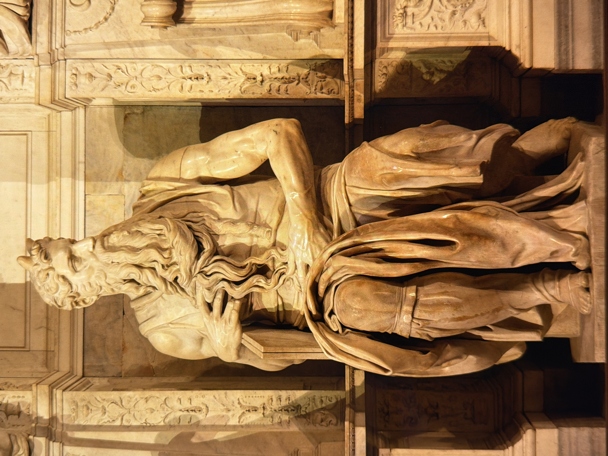 The image is a close-up, eye-level, full-body shot of Michelangelo's sculpture of Moses. The sculpture is carved from marble and depicts Moses seated, with a long, flowing beard and hair, and two small horns protruding from his forehead. He is draped in robes, with his left arm resting on his knee and his right hand tucked into his robe. The sculpture is set within an ornate marble niche, which features carved columns and decorative friezes. The lighting is dramatic, with strong highlights and shadows that emphasize the texture and musculature of the sculpture. The background is a plain, textured wall. There are no people, animals, or other elements in the image apart from the sculpture and its architectural setting. The location is not in Kyiv, Ukraine; this sculpture is located in the Basilica di San Pietro in Vincoli in Rome, Italy.
