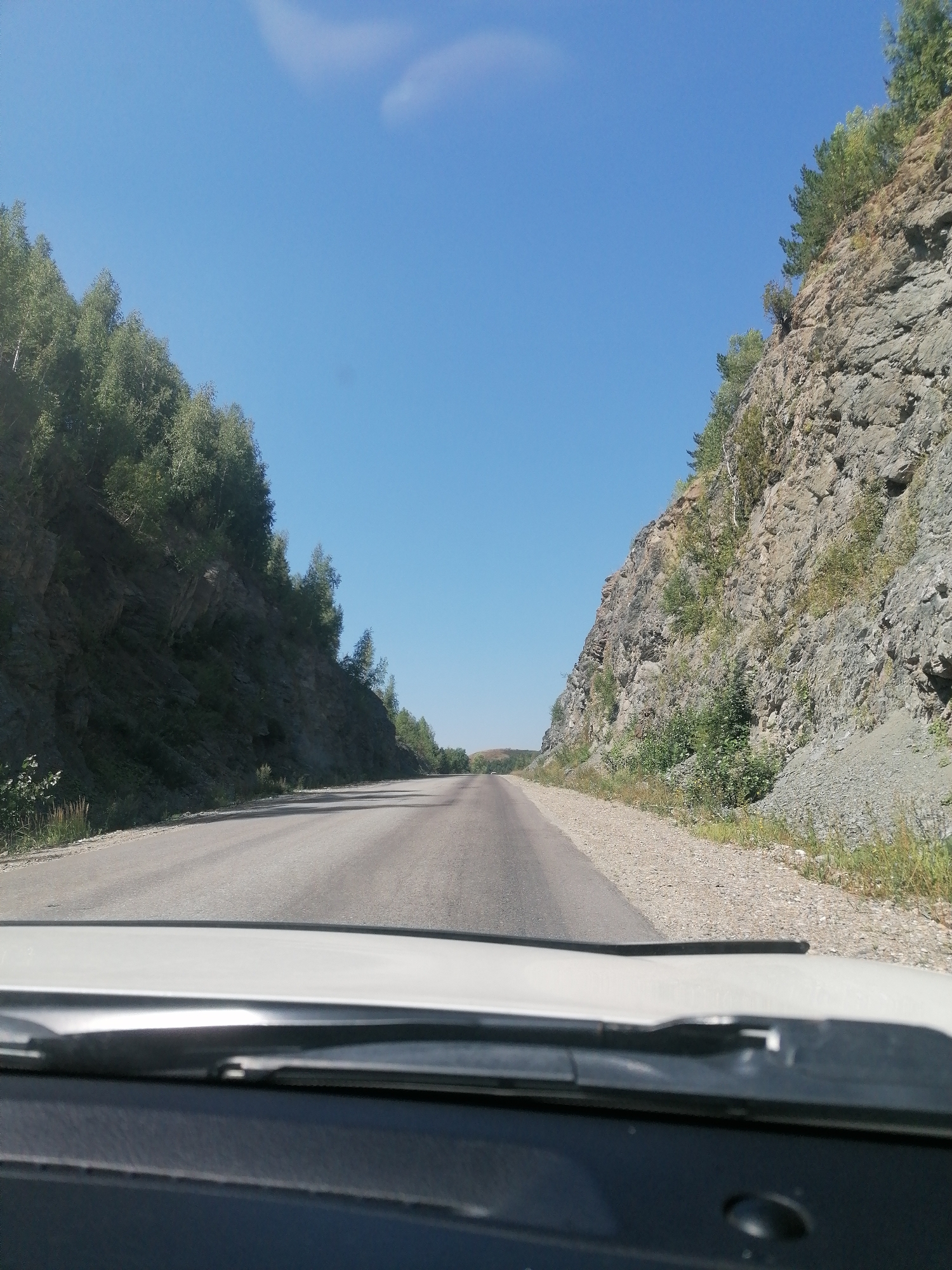 The image shows a view from inside a vehicle, looking forward through the windshield. A paved road stretches ahead, bordered by steep, rocky cliffs on both sides. The cliffs are covered with sparse vegetation, including trees and shrubs. The sky is a clear blue with a few wispy clouds. The road appears to be in a rural or mountainous area. The location is identified as Tarkhanka rural district, Kazakhstan. The time of day appears to be daytime, with bright sunlight casting shadows on the road. There are no people or other vehicles visible in the immediate vicinity. The vehicle's dashboard is partially visible in the foreground, indicating the perspective is from the driver's seat.