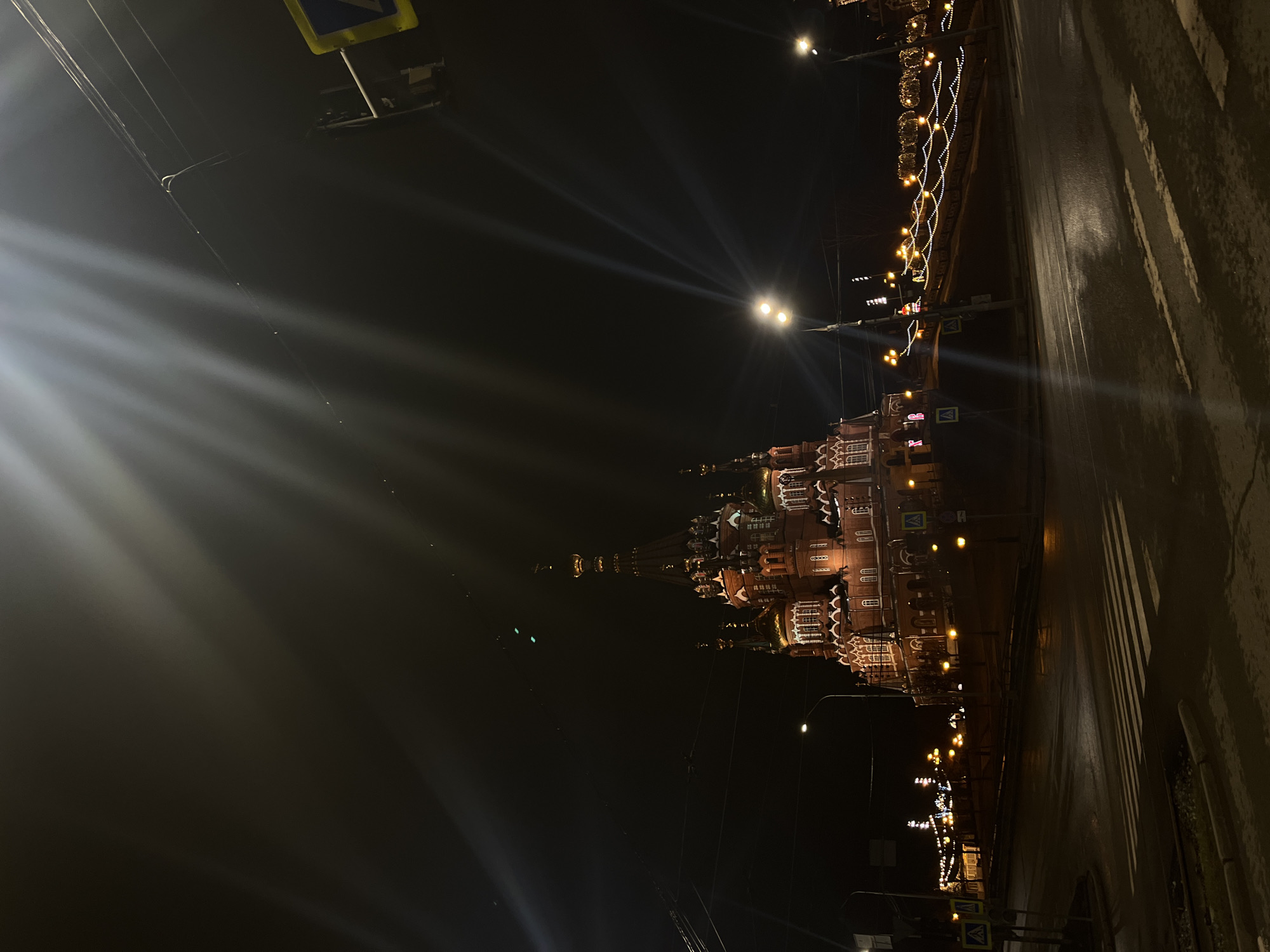 The image captures a nighttime view of the cityscape in Izhevsk, Russia, with the prominent St. Michael's Cathedral illuminated against the dark sky. The cathedral's detailed architecture is visible, accented by decorative lights. Streetlights and holiday decorations along the roadside also add to the illumination. To the right, a wet street is marked with a zebra crossing. In the background, power lines are strung across the sky. Signage is also seen near the pedestrian crossing.