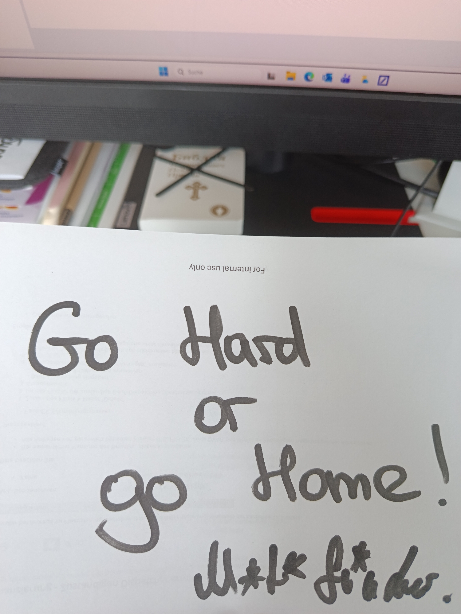 The image captures a desk scene from an angled, slightly elevated perspective, likely in an office or home office environment in Varna, Bulgaria. In the foreground, a white piece of paper is prominently displayed, featuring bold, handwritten text in a dark gray or black marker. The main message reads: "Go Hard or go Home!". Below this motivational phrase, another smaller, handwritten line is visible, stylized with asterisks to resemble an expletive, specifically "M*t* f**k**s." At the very top of the paper, rotated upside down, the text "For internal use only" is printed.
The paper rests on a dark surface, which appears to be a desk. Behind the paper, various desk items are partially visible, including a stack of papers or folders, a distinctive white object with a gold cross symbol and what looks like clock hands, and a red pen or marker.
In the background, a computer monitor screen is visible, displaying parts of a digital interface. The Windows taskbar is discernable at the bottom of the screen, showing the Windows logo, a search icon, and several application icons such as Microsoft Edge and File Explorer. The search bar clearly displays "Q Suche," indicating a German-language operating system. The monitor's screen itself appears to be showing a light-colored background, possibly a document or web page.
The overall scene conveys a sense of a workspace, with the handwritten note adding a personal, informal, and highly motivational or assertive tone. No specific time of day or weather is discernible, but the indoor setting is clearly established.