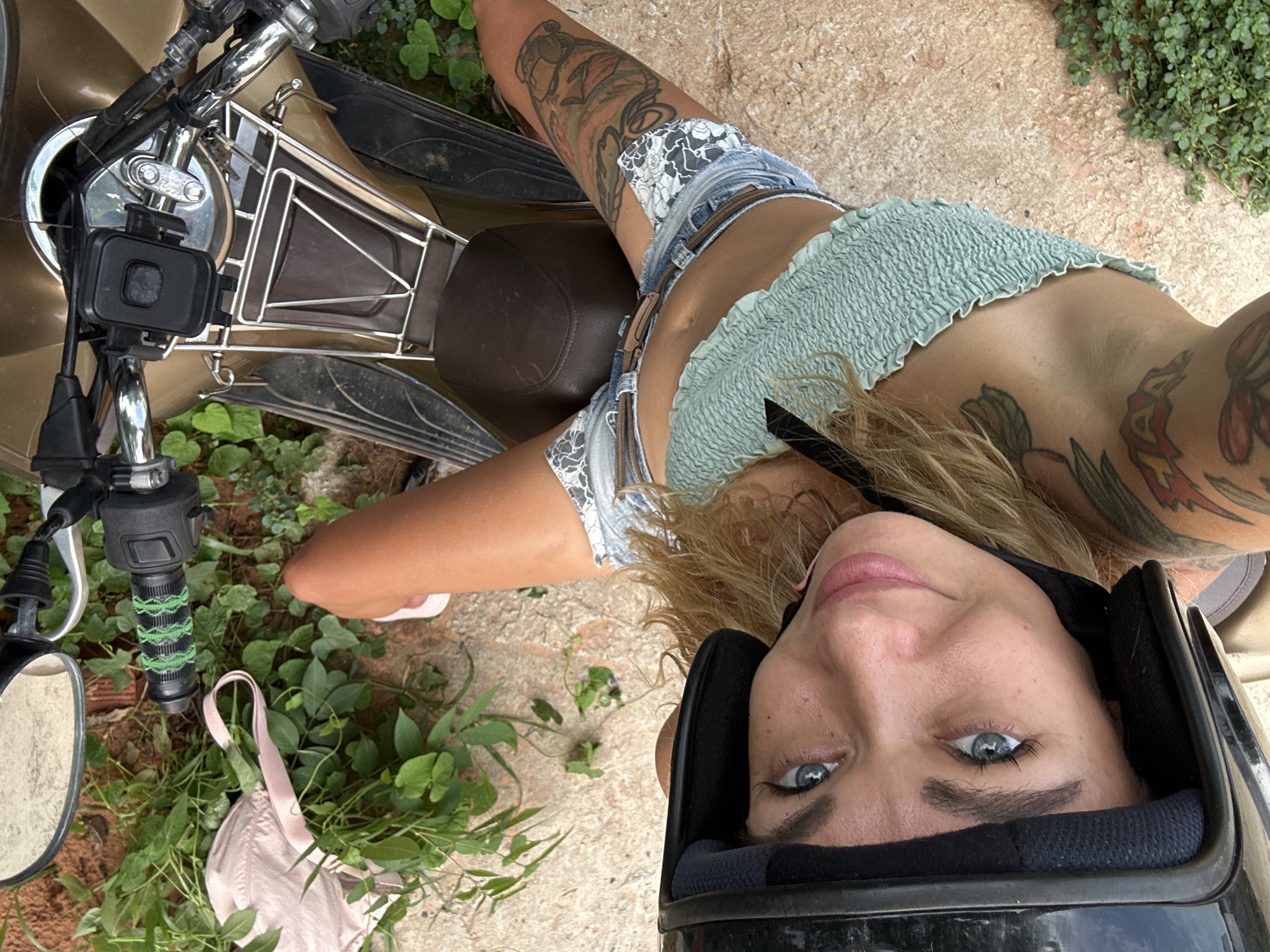 This rotated top-down selfie captures a female subject looking directly at the camera while situated on or next to a light brown scooter in Mũi Né, Vietnam. The subject, a woman with long blonde hair, is wearing a black helmet that frames her face, highlighting her striking blue eyes and natural pink lips. Her expression appears calm and relaxed, with a hint of a smile. She is dressed in a light mint green smocked crop top, denim shorts with white lace or patterned trim, and a brown belt. Her arms and legs are visibly adorned with numerous colorful and intricate tattoos. One pink flip-flop is visible on her right foot.
The scooter, a prominent object in the foreground and midground, is light brown or gold. It features black handlebars with a phone mount attached, a chrome or silver metal front rack, and a brown seat. A side mirror is visible on the left. Lying on the ground near the scooter's handlebars, amidst lush green foliage, is a light pink bra. The ground itself is light-colored and rough, possibly concrete or packed dirt. The setting is clearly outdoors during daytime, indicated by the bright, even lighting and the presence of abundant green plants, consistent with a tropical location. The woman's pose suggests she is either resting on the scooter or taking a self-portrait from a relaxed, reclined position. No discernible text is visible in the image. The overall impression is one of a casual, leisurely moment during a trip.