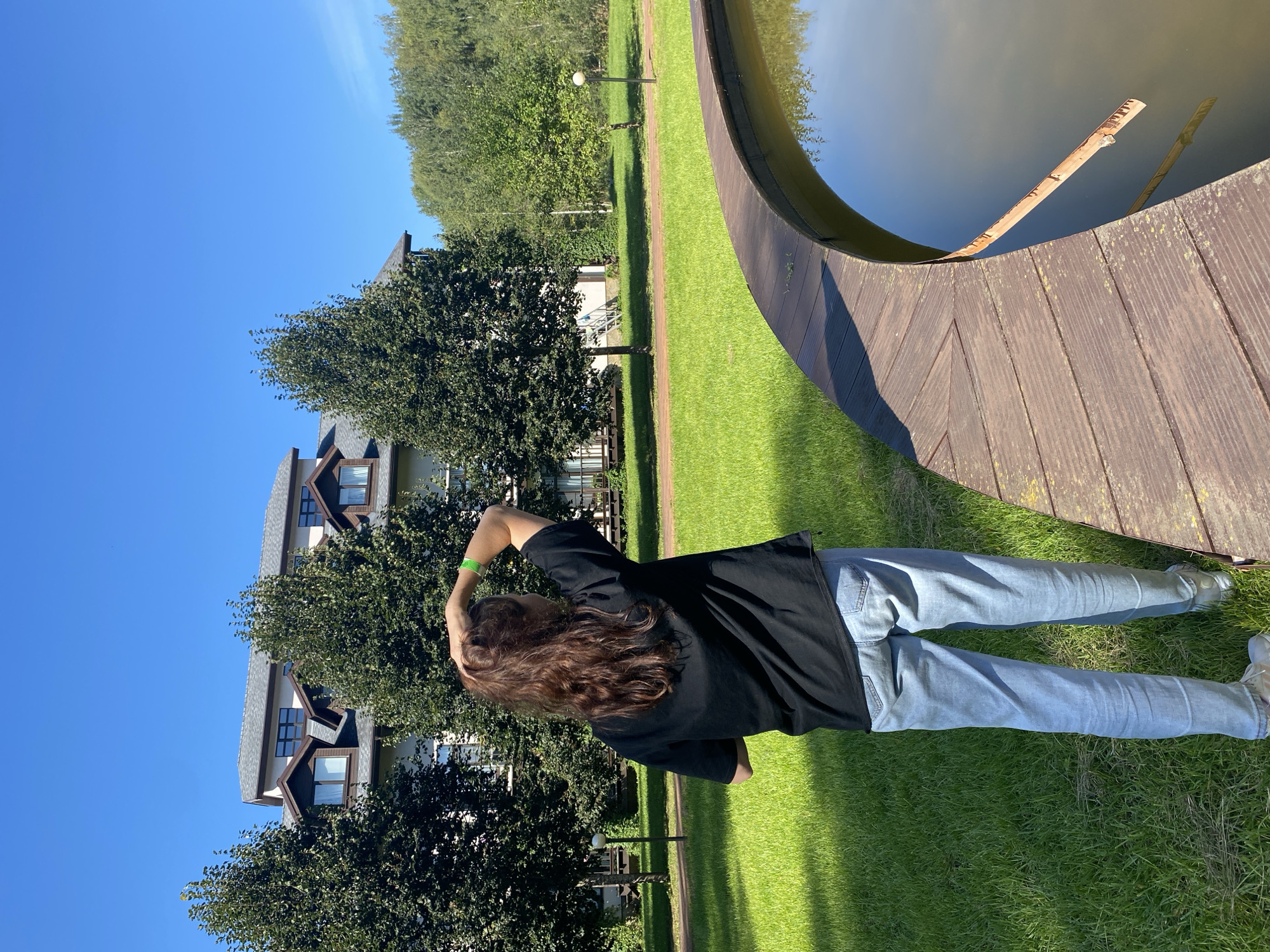 A person with long, brown hair stands with their back to the camera, facing away from a building and towards a pond. They are wearing a black t-shirt and light-wash jeans. Their right arm is raised, with their hand resting on the back of their head. The person is standing on a curved wooden walkway that borders a murky pond. Behind the person, a large, multi-story building with a dark roof and dormer windows is visible, partially obscured by several trees. The building is set back from the walkway and surrounded by a green lawn. Further in the background, more trees and a clear blue sky can be seen. The scene appears to be outdoors, possibly in a park or resort area in Chulis, Russia, on a sunny day.