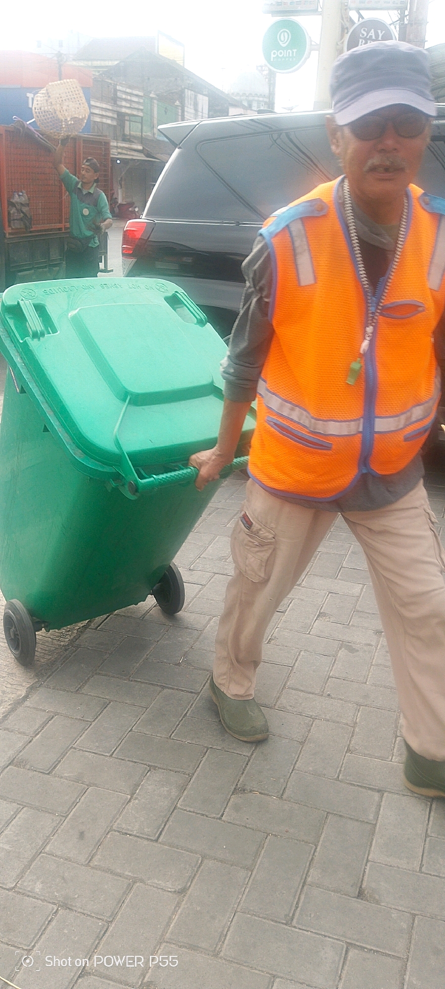 A man wearing an orange safety vest and a blue cap is pulling a green trash bin with wheels on a paved sidewalk. He is wearing tan pants and green boots. In the background, another man in a green shirt is visible near a red truck, holding a woven basket. A black SUV is parked behind the trash bin. The environment appears to be an outdoor urban setting with buildings and signs in the distance, including a "Point Coffee" sign. The image is taken from a slightly low angle, focusing on the man with the trash bin.