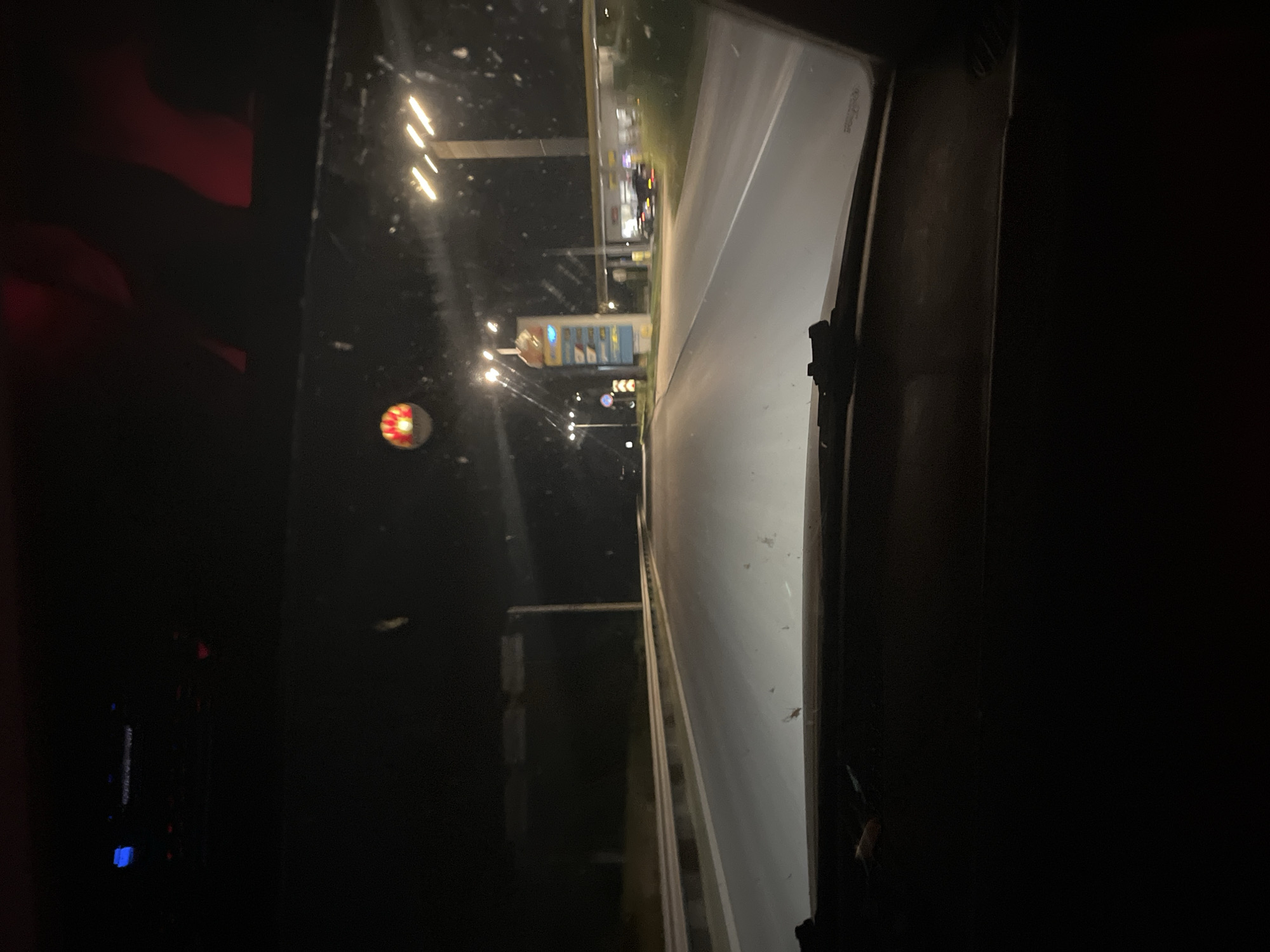 The image captures a nighttime scene from what appears to be the interior of a vehicle, looking out through a windshield or window. The perspective is slightly angled, suggesting movement.
The main subject is the road ahead, illuminated by streetlights and the lights of a building that resembles a gas station or convenience store. The building has prominent signage, including what looks like a "Shell" logo and price displays. Multiple lights are visible on poles and on the building itself, casting a glow on the wet asphalt of the road, indicating either recent rain or dew.
To the left of the road, there are dark areas with hints of red light, possibly from interior lights of the vehicle or reflections. A circular, red and white object, possibly a decorative light or a warning signal, is visible in the lower left quadrant. Further back, more lights suggest the presence of other structures or vehicles.
The overall atmosphere is dark and nocturnal, with artificial lights being the primary source of illumination. There are no people clearly visible, but the scene implies human activity through the presence of the road and the commercial establishment. The time of day is unequivocally night. There are no explicit weather cues beyond the potential wetness of the road.
The location context of Yasen, Bulgaria, provides a geographical anchor, but no specific visual cues within the image directly confirm this location. The visible text, "Shell," is a brand name common globally. The scene evokes a sense of travel or a stop during a nighttime journey.