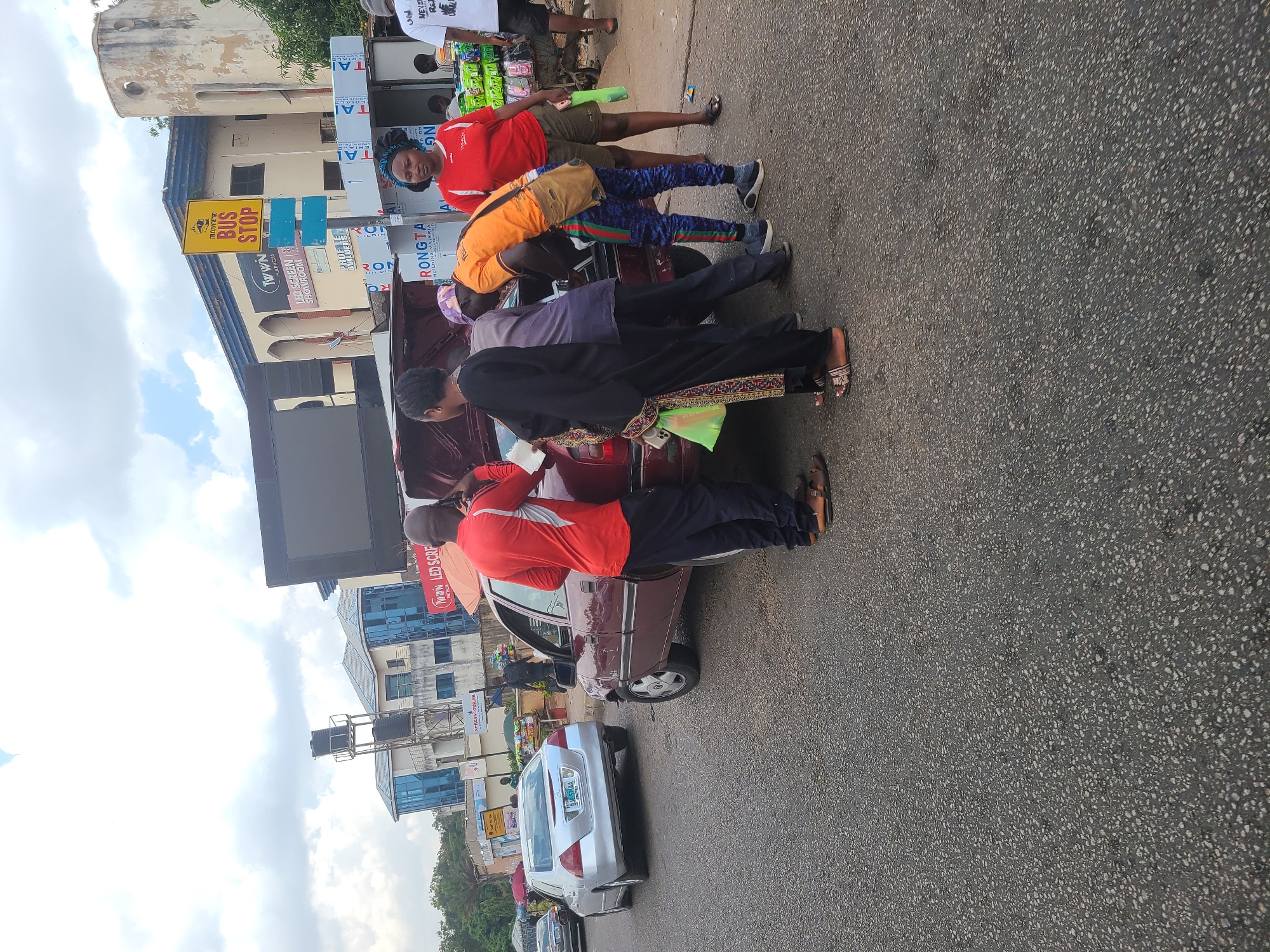 The image depicts a street scene in Jos, Nigeria, featuring a burgundy-colored car with its trunk open. Four people are positioned around the open trunk, appearing to interact with the contents or each other. One person wears a red shirt and leans into the trunk, another is dressed in orange, and a third in black attire. A fourth person wears a red shirt and black pants. To the left of the car is a silver sedan, followed by other vehicles further down the road. A "Bus Stop" sign is prominently displayed alongside signs for "NAAL MULTIMEDIA LED SCREEN SHOWROOM" and "RONGTA BUILDING MATERIALS." The background includes buildings, trees, and a cloudy sky. The road surface is asphalt.
