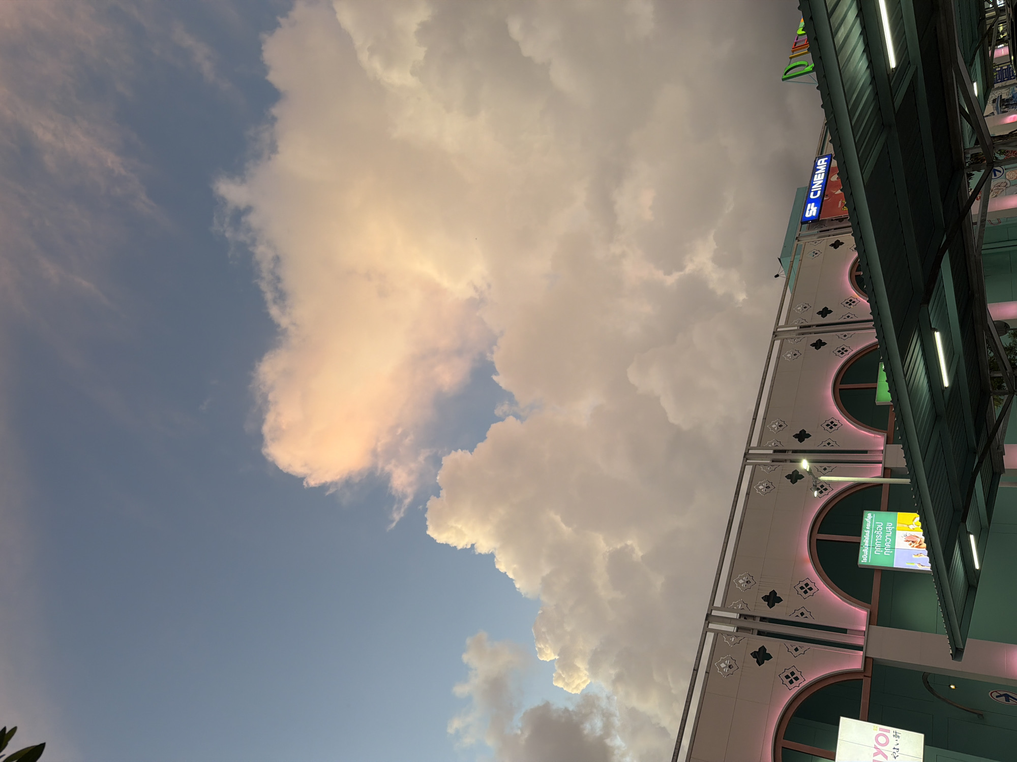 This is a vertical, eye-level, outdoor shot of the sky and the upper portion of a building's facade in Si Sunthon, Thailand. The sky is a blend of blue and soft orange, with large, fluffy clouds illuminated by the sun. The clouds are the dominant feature in the upper half of the image. Below the sky is the exterior of a building with arched windows highlighted by pink neon lighting. The facade is adorned with decorative patterns. A dark green awning covers the foreground, with fluorescent lights visible underneath. Signs are affixed to the building; one prominent blue neon sign reads "SF CINEMA." The scene appears to be captured during either dawn or dusk, given the lighting conditions. There are no people visible in the image.
