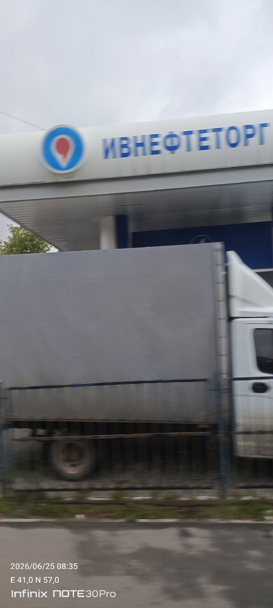 The image captures a motion-blurred view of a gas station canopy with the brand "ИВНЕФТЕТОРГ" (IVNEFTETORG) displayed prominently in blue lettering. A company logo, a red pin on a blue circle, is visible to the left of the text. Below the canopy, a large, grey-covered truck is partially visible, also affected by motion blur. The truck appears to be on a road or at a service area. The timestamp and GPS coordinates "2026/06/25 08:35 E 41,0 N 57,0" are overlaid on the bottom left of the image, along with the device information "Infinix NOTE 30Pro". The background is a hazy, overcast sky. The location appears to be on the outskirts of Ivanovo, Russia, based on the GPS coordinates provided, though the specific city environment is not clearly discernible due to the motion blur and framing.