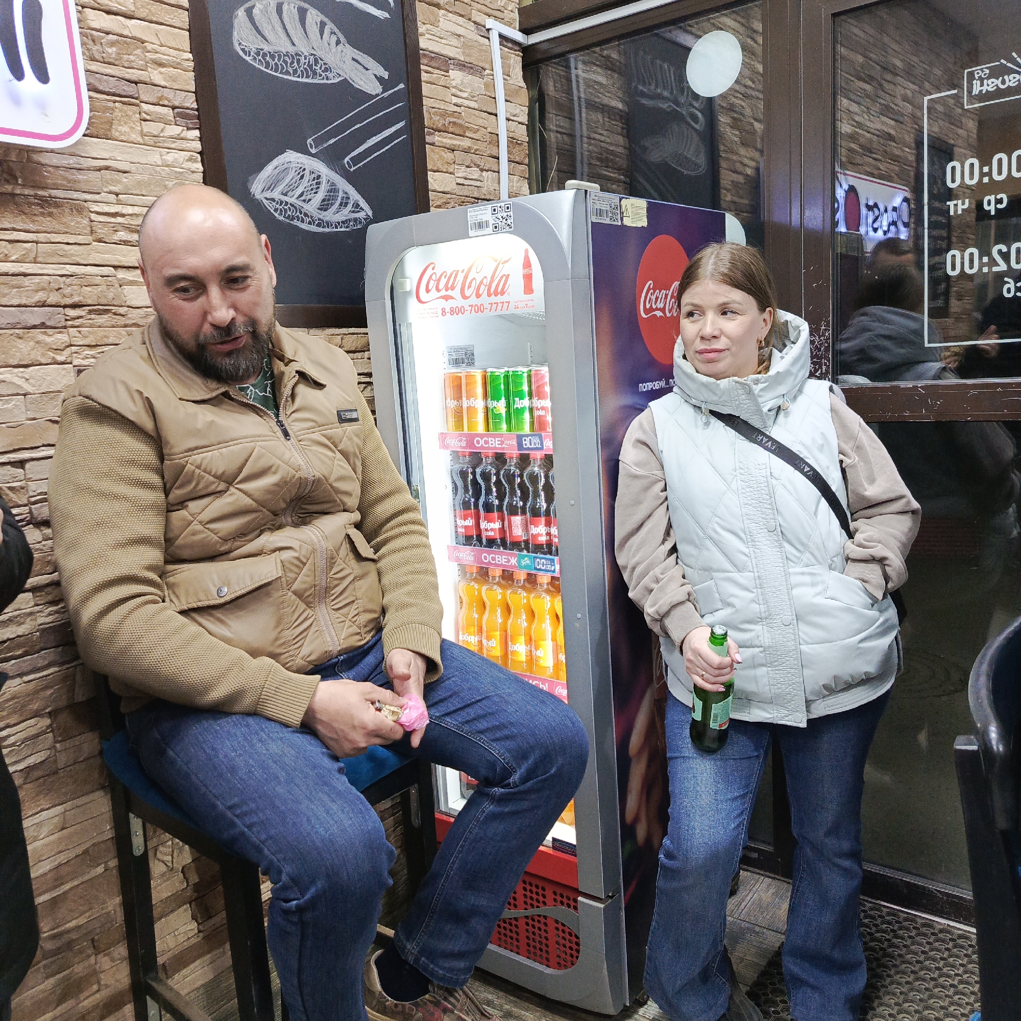 The image shows a man and a woman standing next to a Coca-Cola vending machine in what appears to be a storefront. The man is sitting on a stool, wearing a brown quilted jacket and blue jeans, and holding a pink object in his hands. He has a beard and a bald head. The woman is standing next to the vending machine, wearing a light blue vest over a beige hoodie and blue jeans. She is holding a green bottle, possibly beer. The Coca-Cola vending machine is filled with Coca-Cola products, including orange soda and Coke. The vending machine is located in what seems to be a cafe, restaurant, or convenience store. The background includes a brick wall, a blackboard with sushi illustrations, and a window with reflections and signage. The OCR detected identifies the city as Chaikovsky, Russia. The signage in the window appears to show opening and closing times.