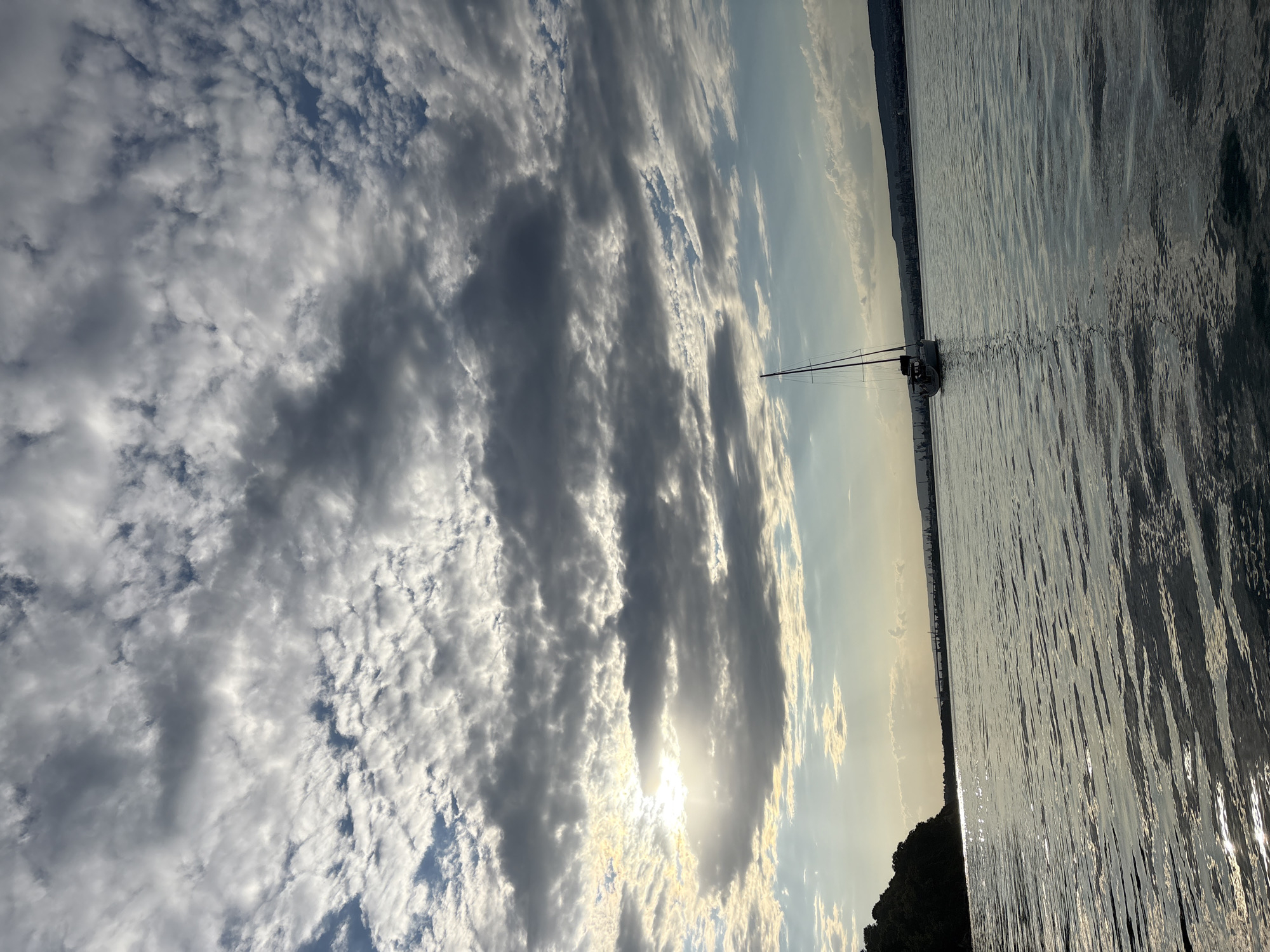 A wide shot captures a sailboat navigating a body of water under a dramatic sky filled with clouds. The sun is partially obscured by clouds, casting a soft light across the scene. The water's surface reflects the sky and sunlight, creating ripples and shimmering patterns. On the left, a tree-covered hill slopes down to the water's edge, hinting at a coastline. In the distance, a hazy cityscape with industrial elements, including cranes, stretches across the horizon. The sailboat, with its mast reaching towards the sky, is the primary subject in the water. The overall impression is one of a serene, yet somewhat moody, maritime landscape.