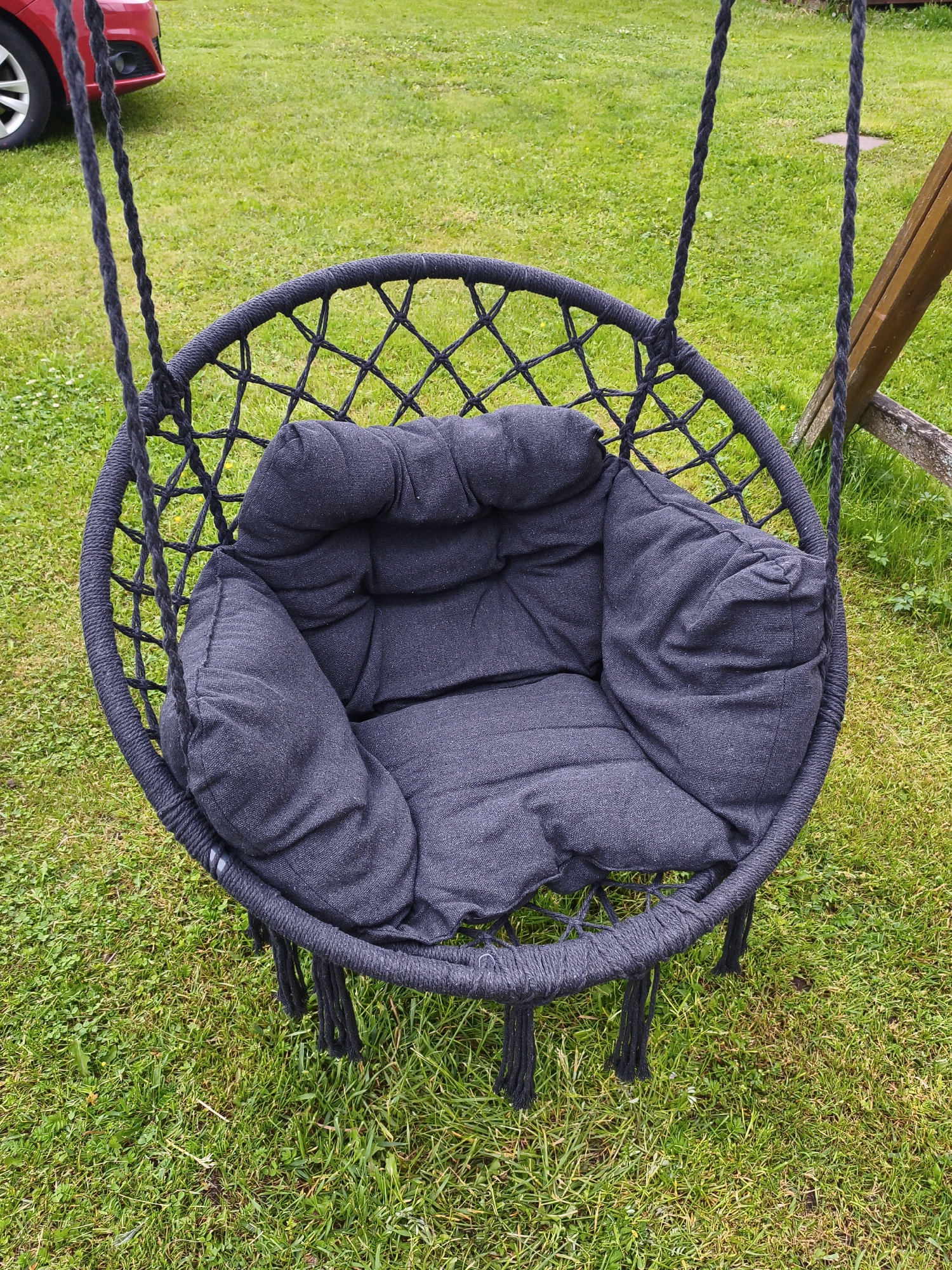 An empty, dark grey fabric cushion sits inside a black macrame hammock chair. The chair hangs from thick black ropes attached to a wooden support structure, partially visible on the right. The chair rests on a well-maintained green lawn. In the background, a portion of a red car is visible on the left side of the frame. The overall scene suggests an outdoor setting, possibly a backyard or park. The location is Brodi, Latvia.