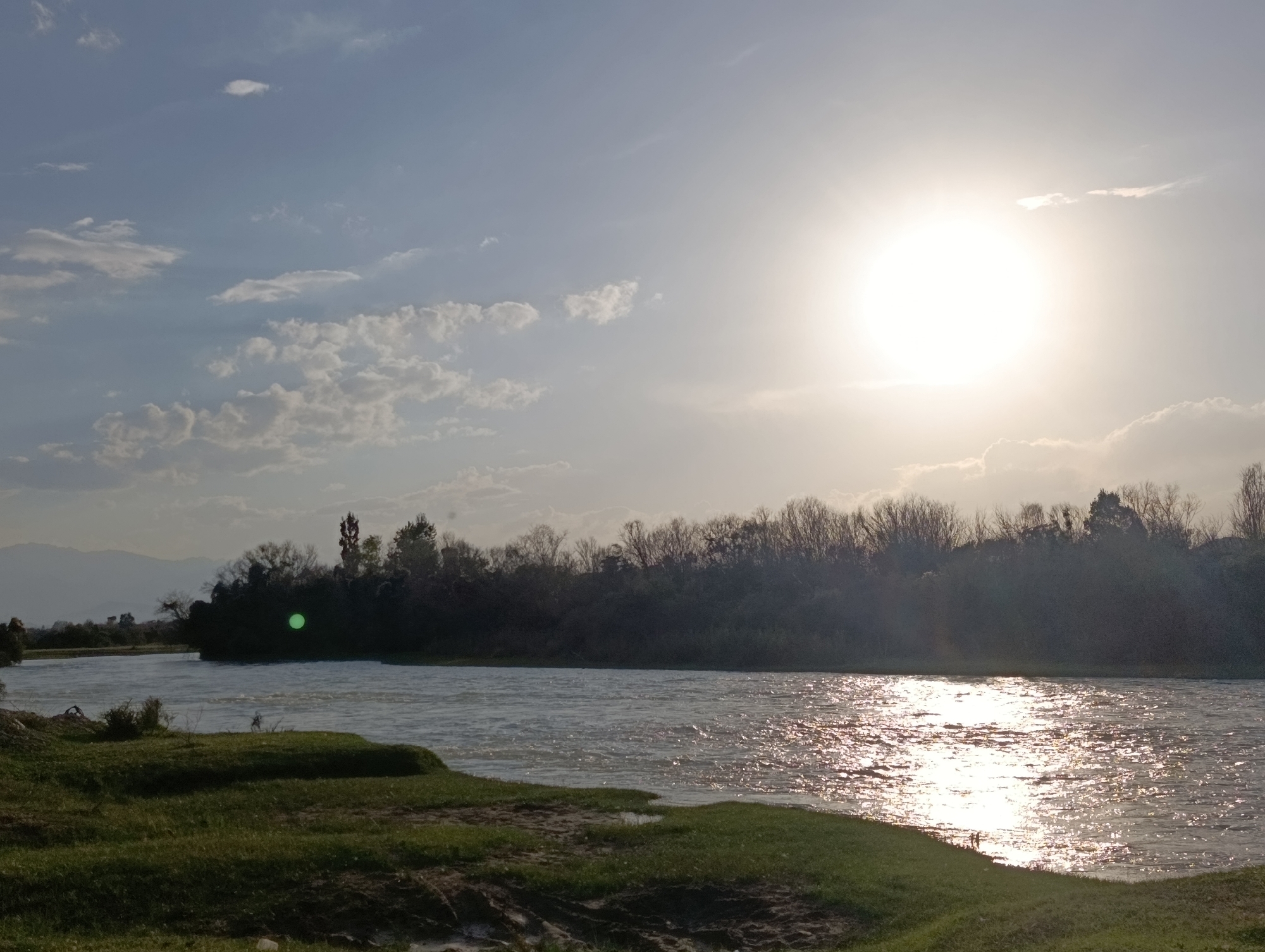 The image depicts a wide, flowing river in Kochkor, Kyrgyzstan. The sun is prominently positioned in the upper right quadrant of the sky, casting a bright glare and creating a shimmering reflection on the water's surface. The riverbanks are lined with lush green grass and some sparse vegetation. In the background, a dense line of trees forms a dark silhouette against the sky. Beyond the trees, faint mountain ranges are visible in the distance. The sky is a pale blue, with scattered clouds that are tinged with the bright light of the sun. There are no people or animals visible in the scene.